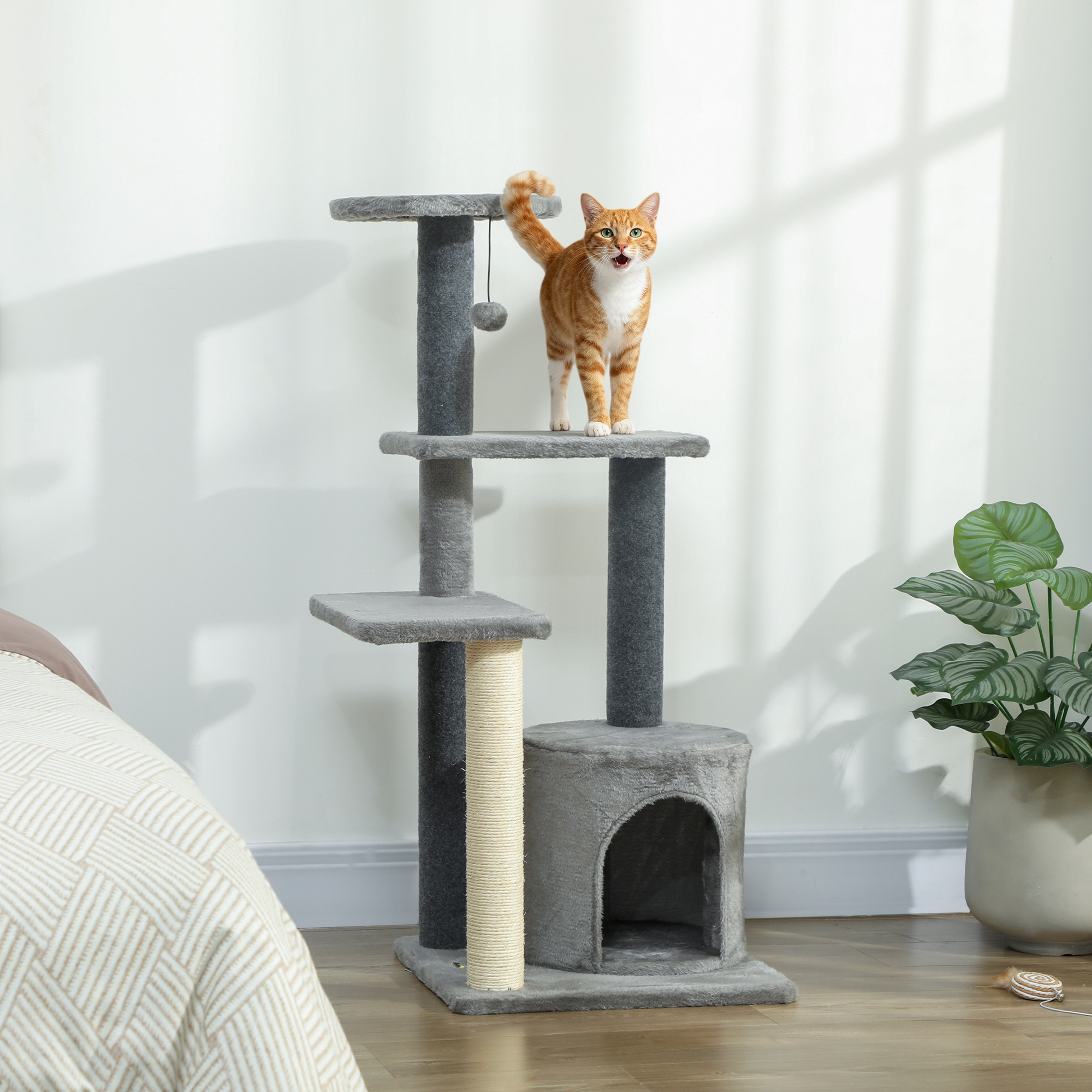 Un gatto soriano arancione si trova su un tiragraffi grigio accanto a un letto e una pianta.