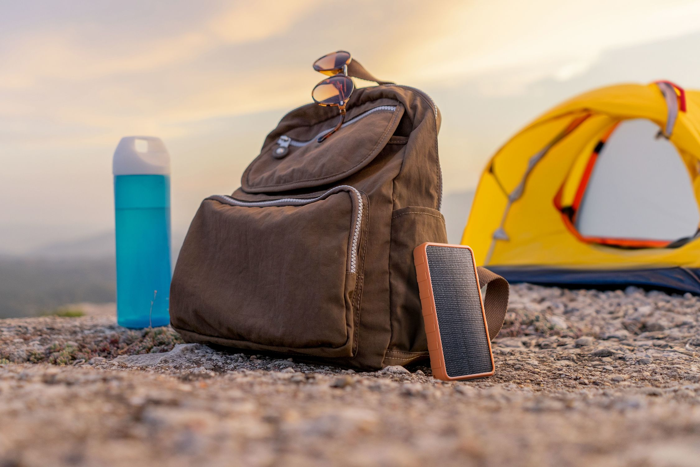 Sac à dos marron, bouteille bleue, lunettes de soleil, enceinte et tente jaune sur terrain rocheux.