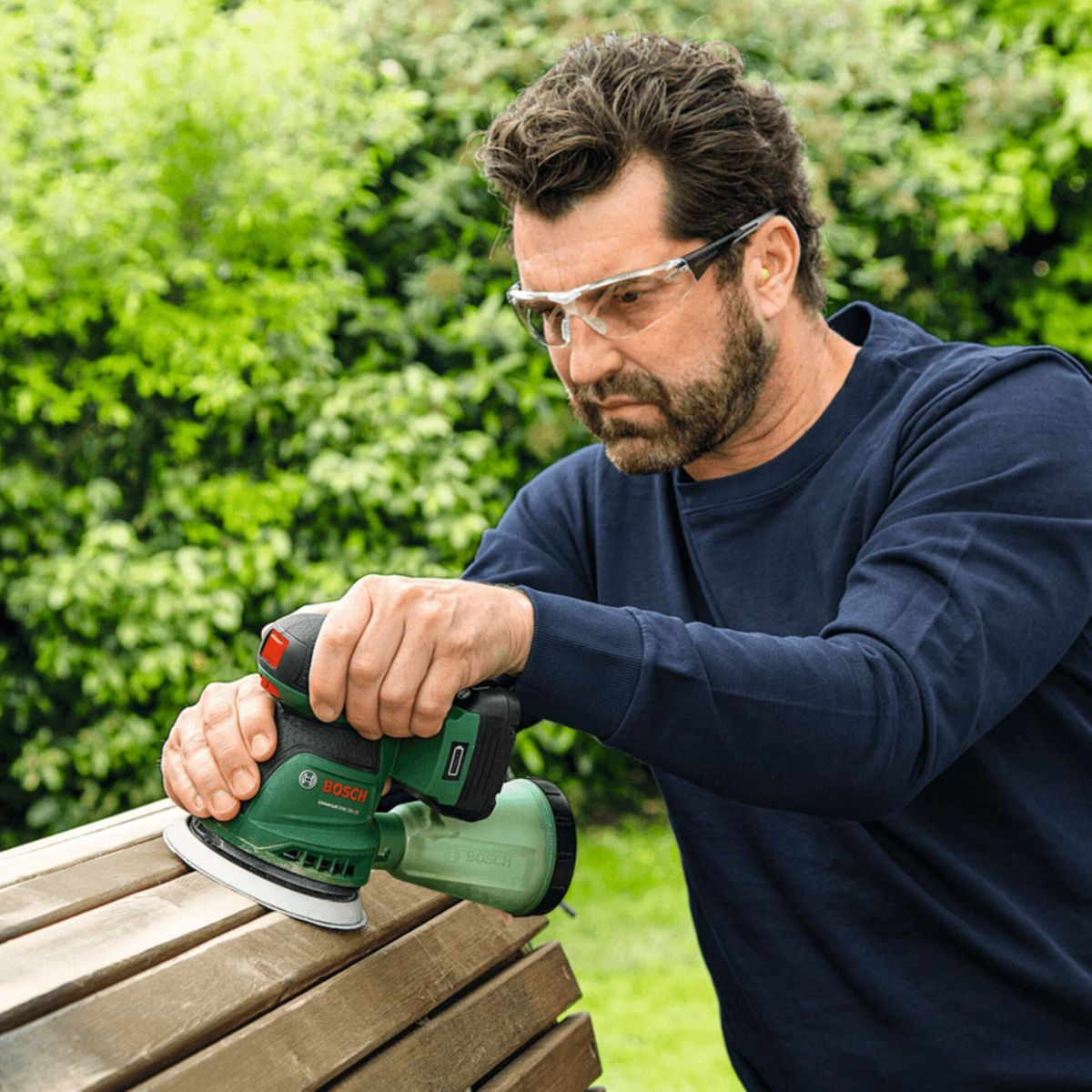 Homme ponçant banc en bois dehors avec ponceuse Bosch verte, portant lunettes.