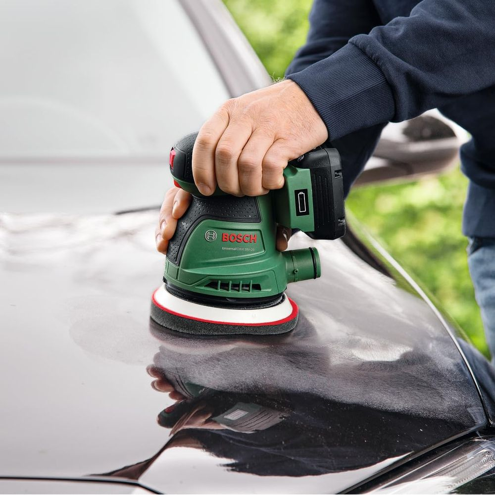 Mains polissant une voiture sombre avec une ponceuse orbitale Bosch verte. Verdure en arrière-plan.