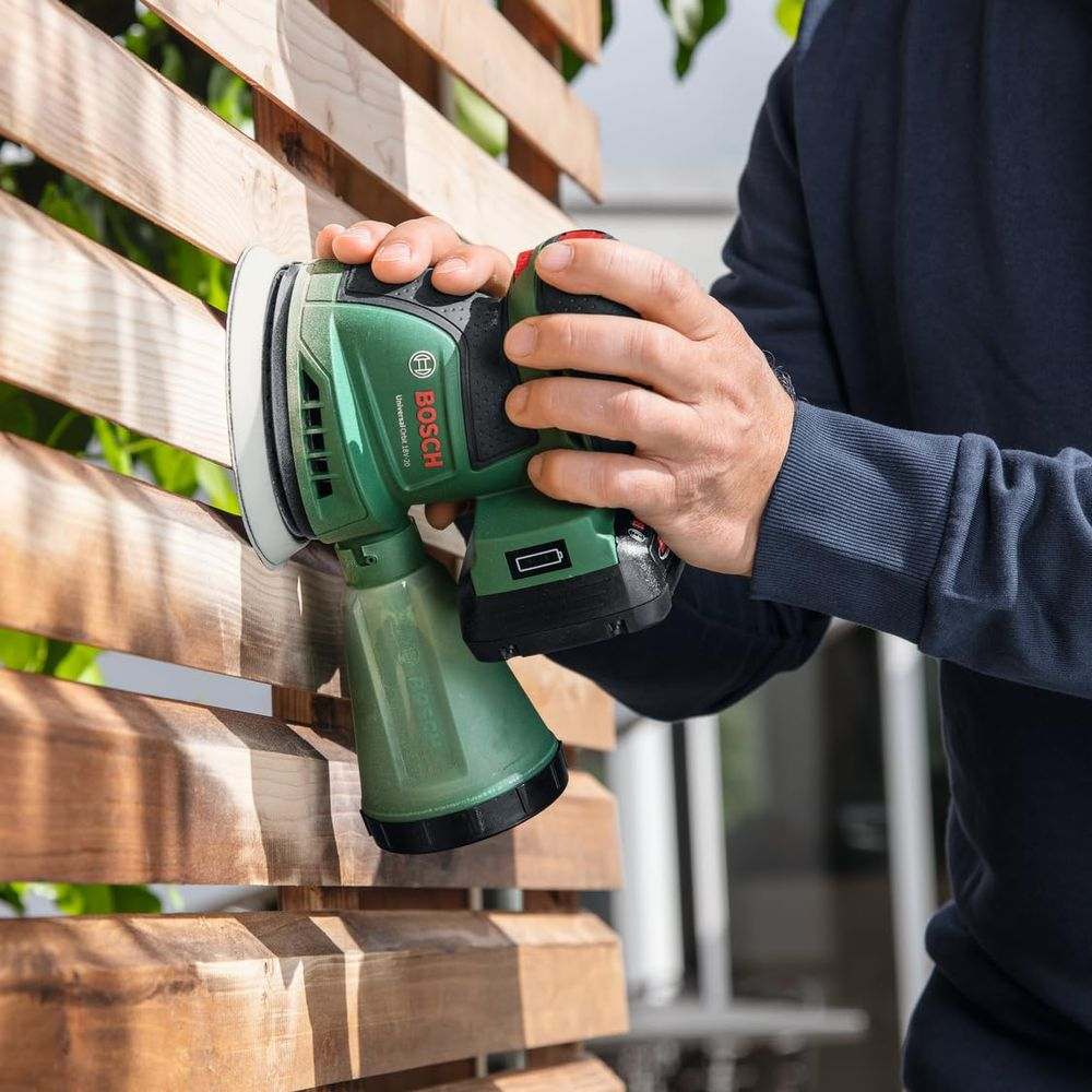 Personne ponçant une clôture en bois avec une ponceuse Bosch verte. Gros plan sur les mains et l'outil.
