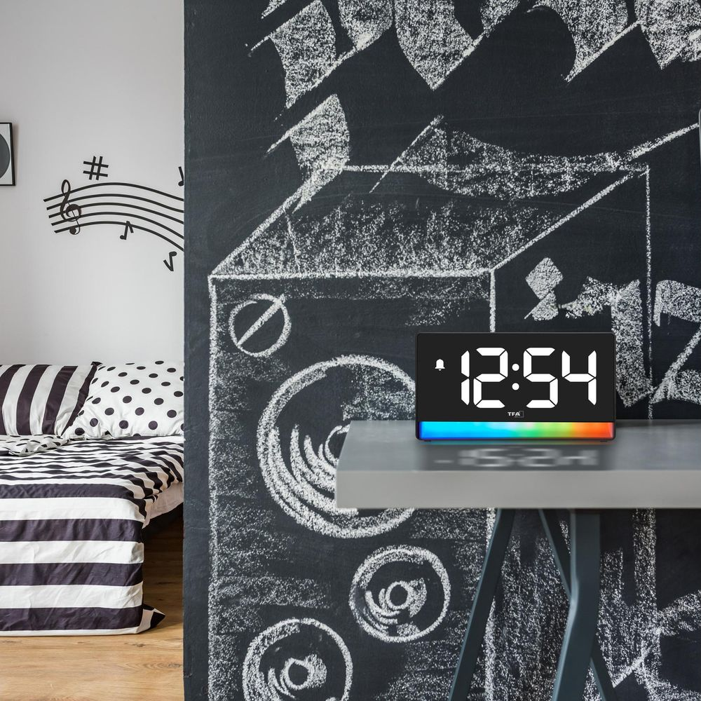 Réveil numérique avec lumière arc-en-ciel sur un bureau devant un tableau noir avec des notes de musique.