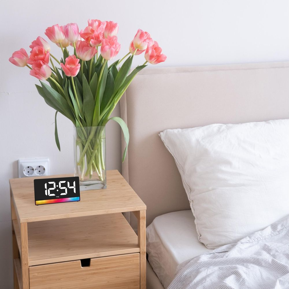 Une table de chevet avec un réveil numérique et des tulipes roses près d'un lit.