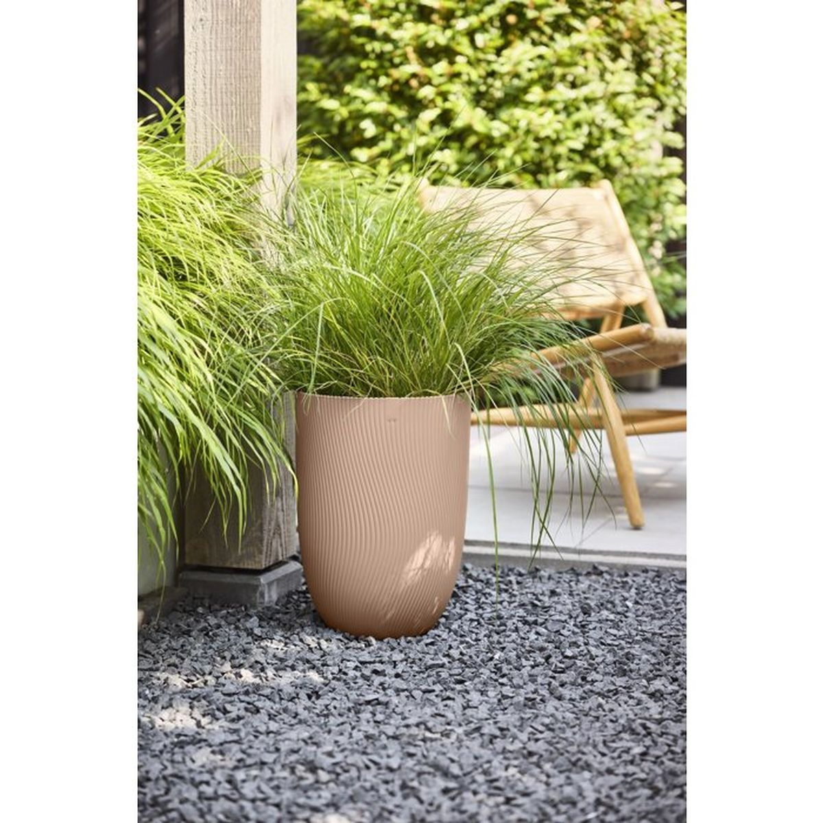 Ein beiger Blumentopf mit Gräsern steht auf grauem Kies neben einem Holzpfosten.