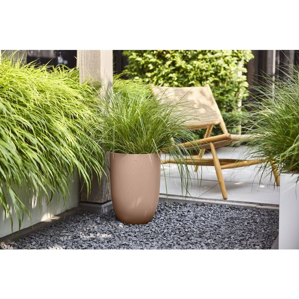 Ein hoher, gerippter, hellbrauner Pflanzkübel mit grünem Gras steht auf dunklem Kies.