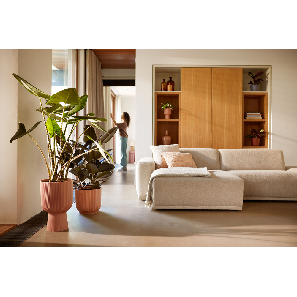 Ein Wohnzimmer mit Sofa, Pflanzen und einer Frau, die am Fenster steht.