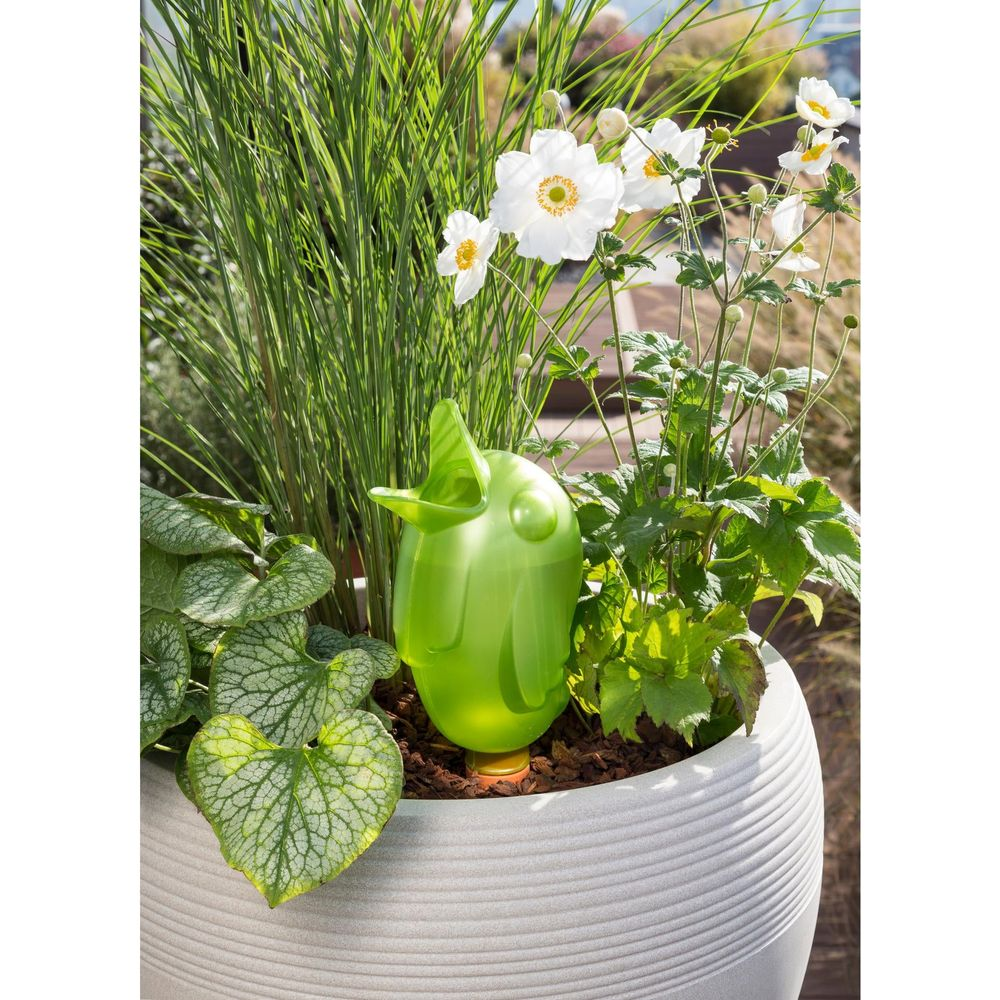Grüner Vogel-Bewässerungsbehälter in einem Topf mit Pflanzen und weißen Blumen.