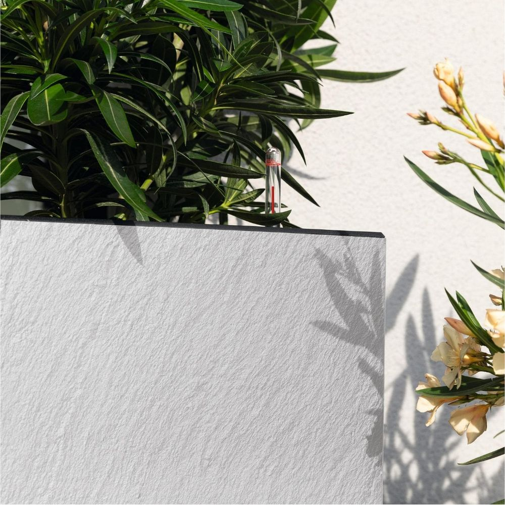 Ein kleines Thermometer mit roter Flüssigkeit steht zwischen grünen Blättern und hellen Blumen.