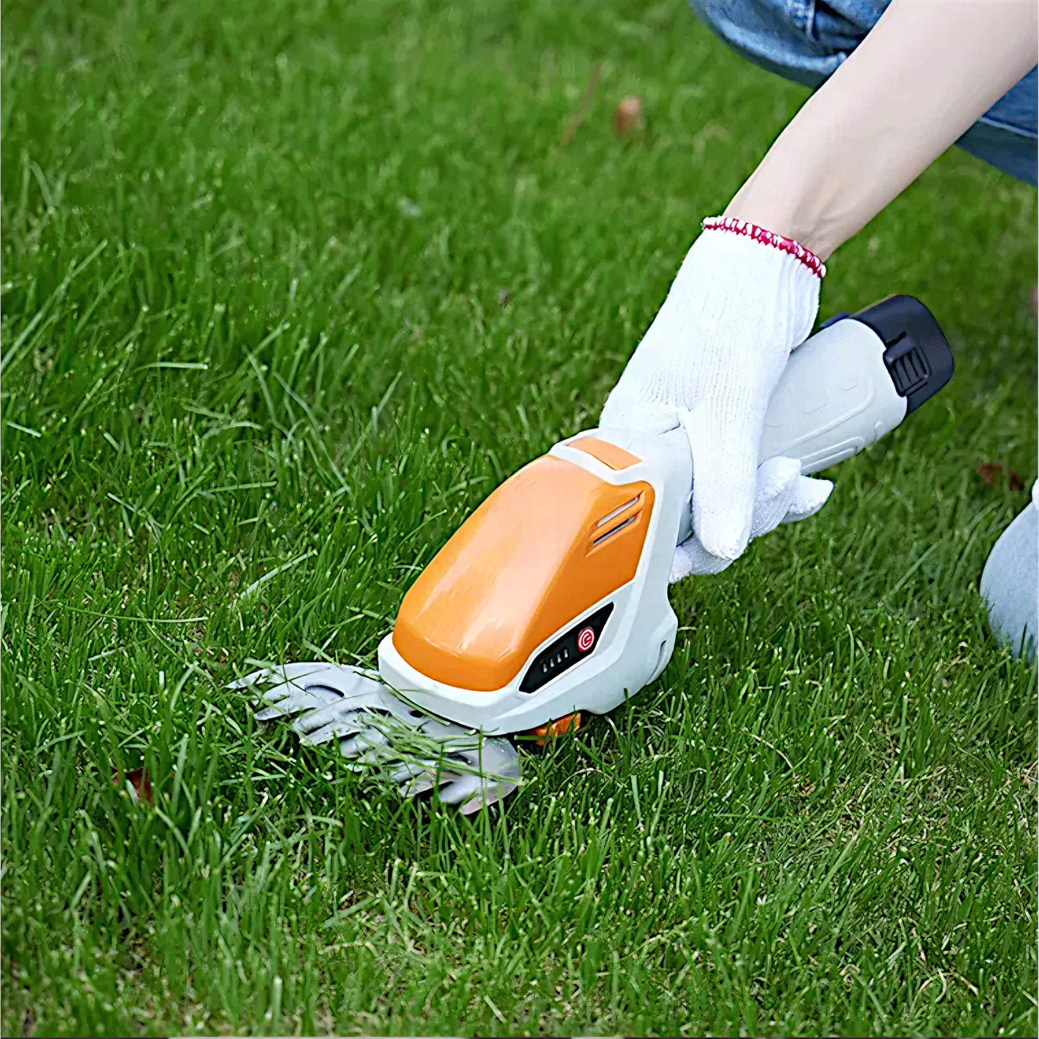 Mano con guante blanco sujetando cortasetos naranja-blanco sobre césped verde.