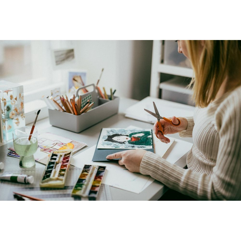 Frau schneidet Kunstwerk mit Schere an einem Schreibtisch mit Kunstbedarf aus.