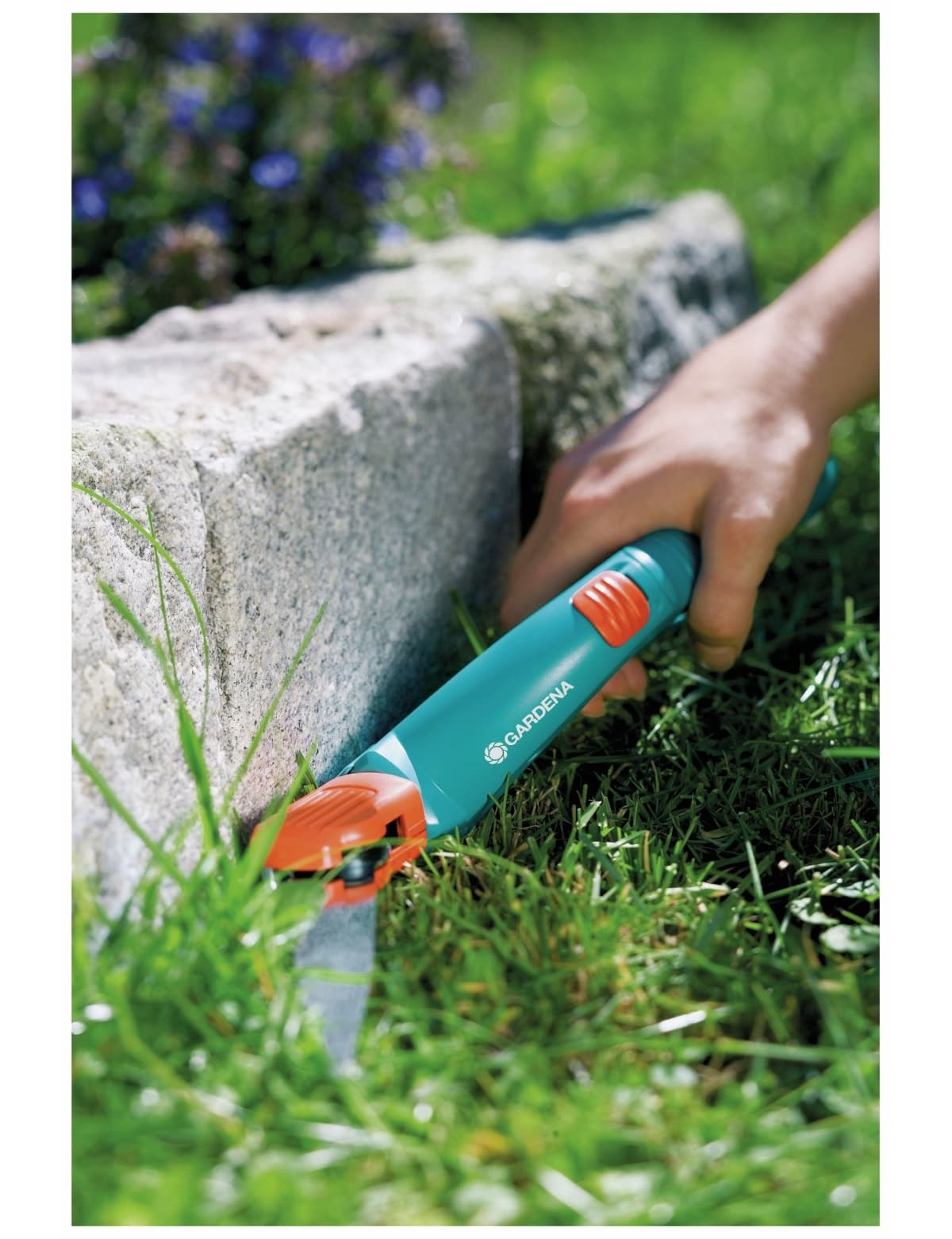 Eine Hand hält einen türkisfarbenen Gartentrimmer mit orangefarbenen Akzenten, der Gras nahe einer Steinbegrenzung schneidet.
