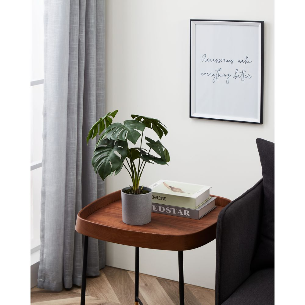 Una pianta di monstera siede su un tavolino di legno accanto a una sedia grigia e tende trasparenti.