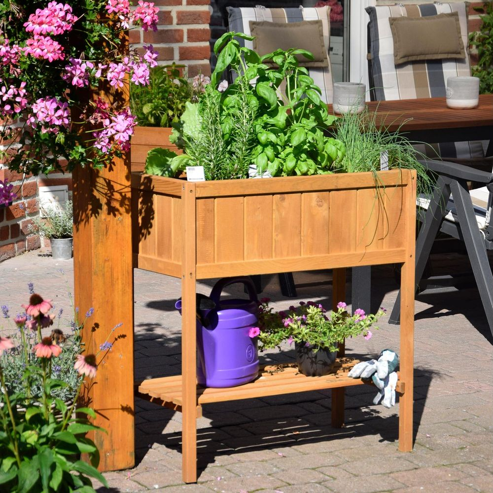 Ein hölzerner Hochbeet mit Kräutern, daneben eine Gießkanne und Blumen.