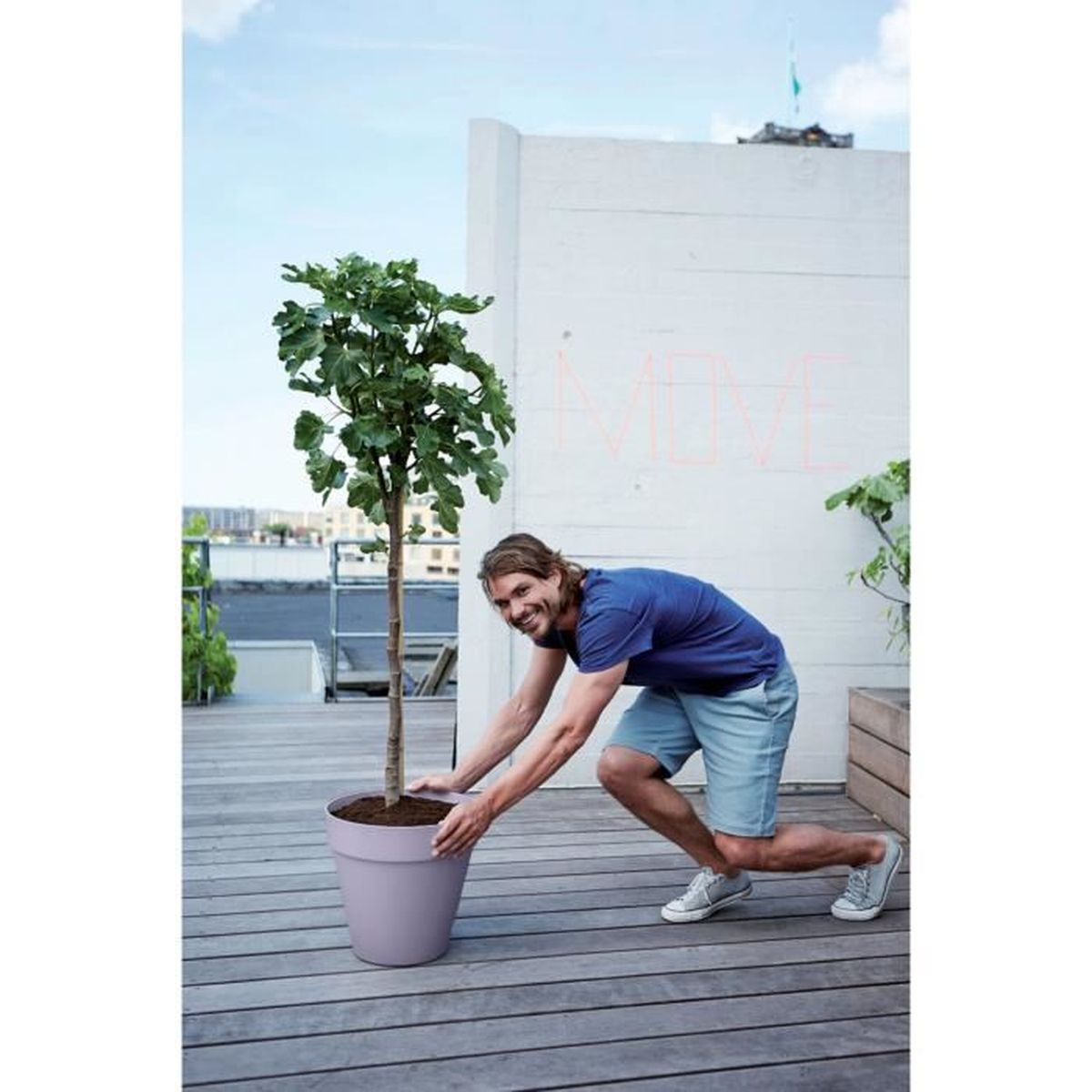 Ein Mann in blauer Kleidung bewegt einen großen Topf mit einem Baum auf einer Holzterrasse.