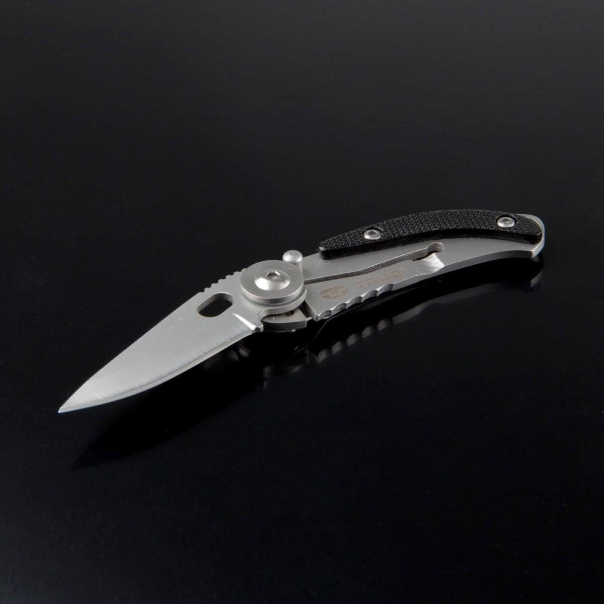 Una navaja plateada con mango negro está sobre una superficie negra reflectante.