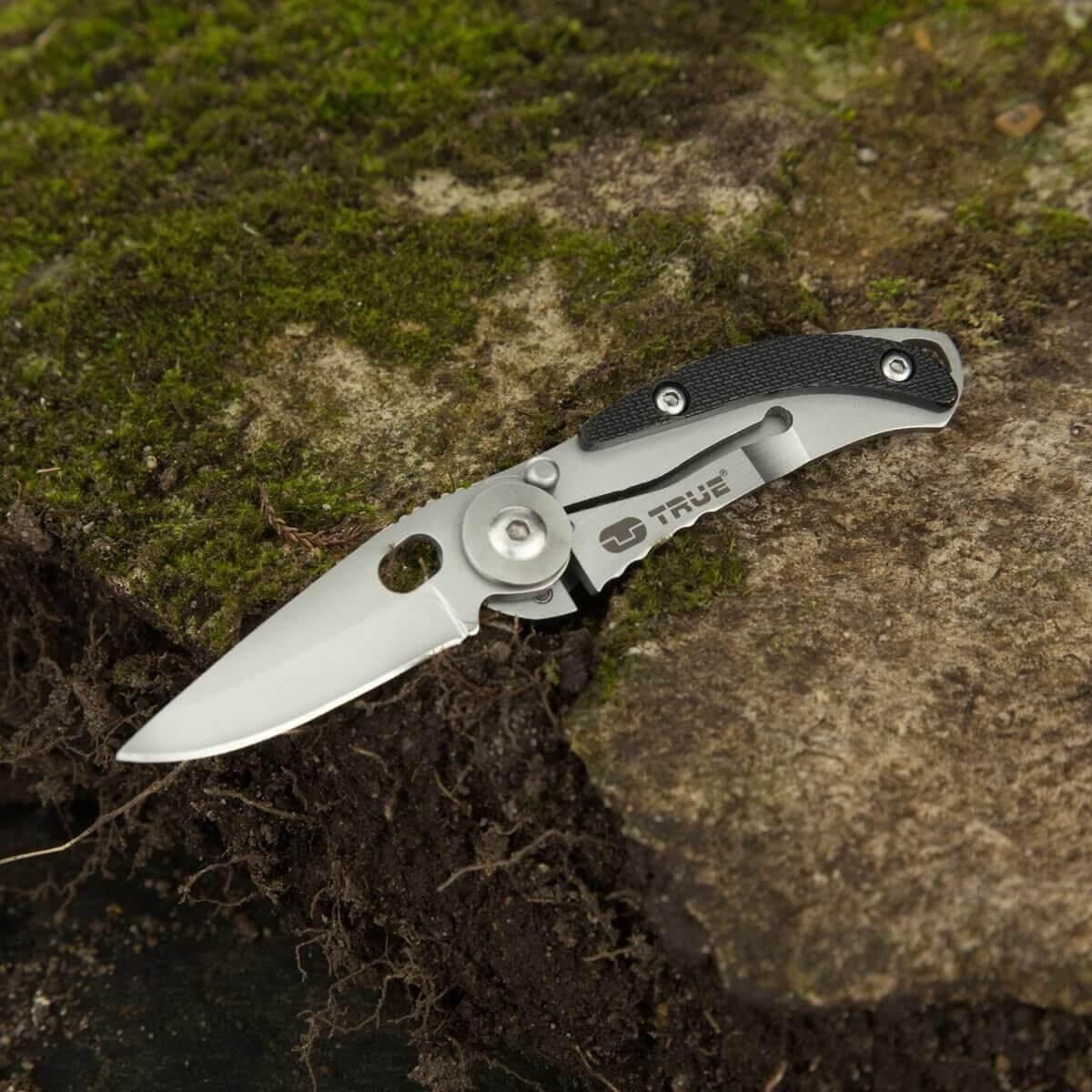 Una navaja plateada con mango negro descansa sobre tierra musgosa y rocas.