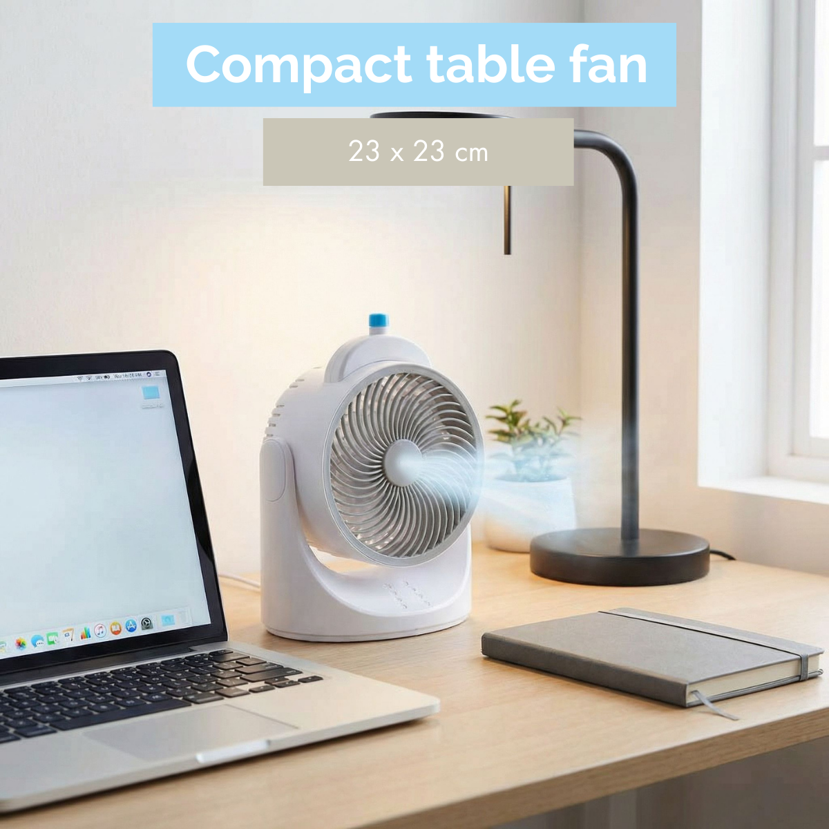 A white compact table fan blows air on a desk with a laptop, lamp, and notebook.