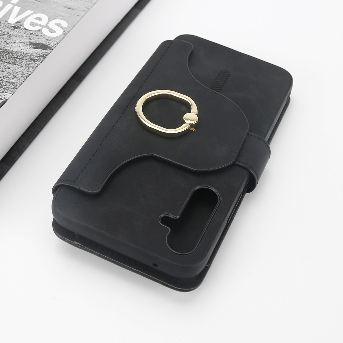 Schwarzes Handy-Etui mit goldenem Ringhalter, neben einem offenen Buch.