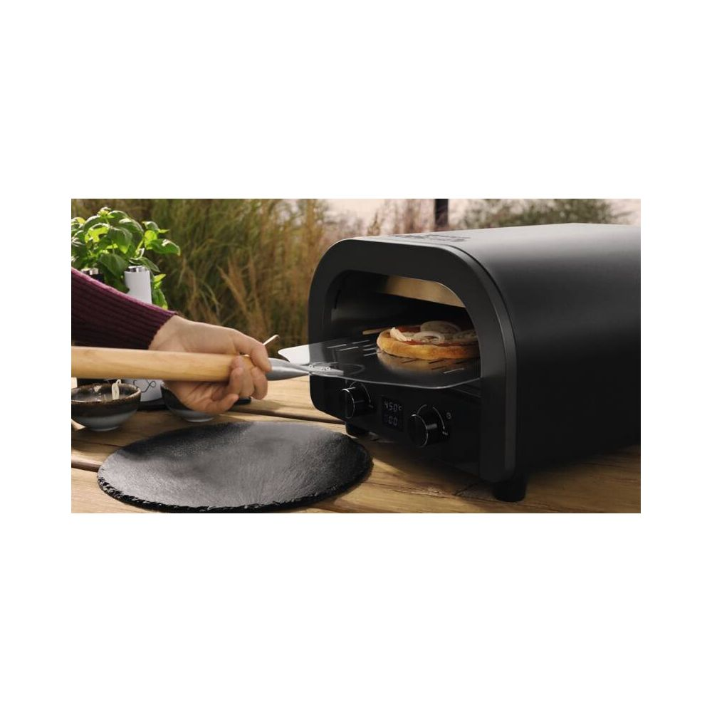 Une main utilise une pelle à pizza pour placer une pizza dans un four extérieur noir sur une table en bois.