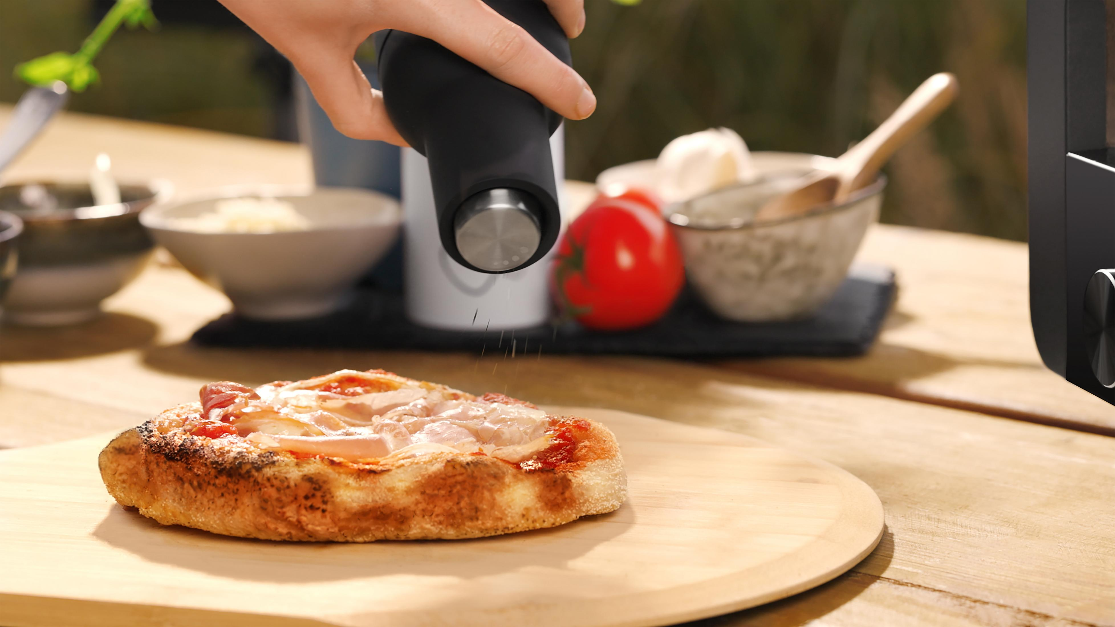 Une main saupoudre des épices sur une petite pizza. Des ingrédients comme des tomates sont à l'arrière-plan.