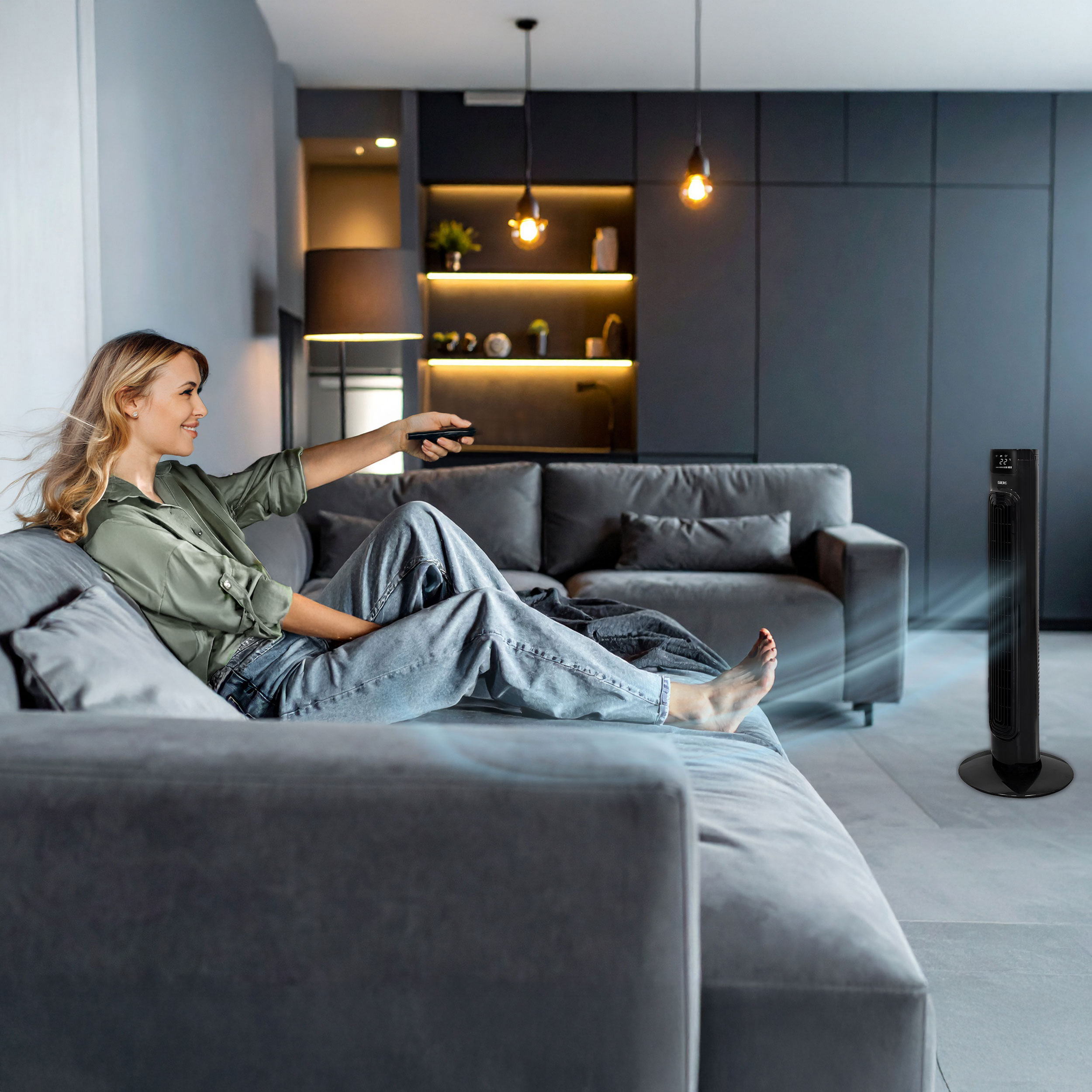 Kobieta na kanapie używa pilota do wentylatora wieżowego rozprowadzającego powietrze.