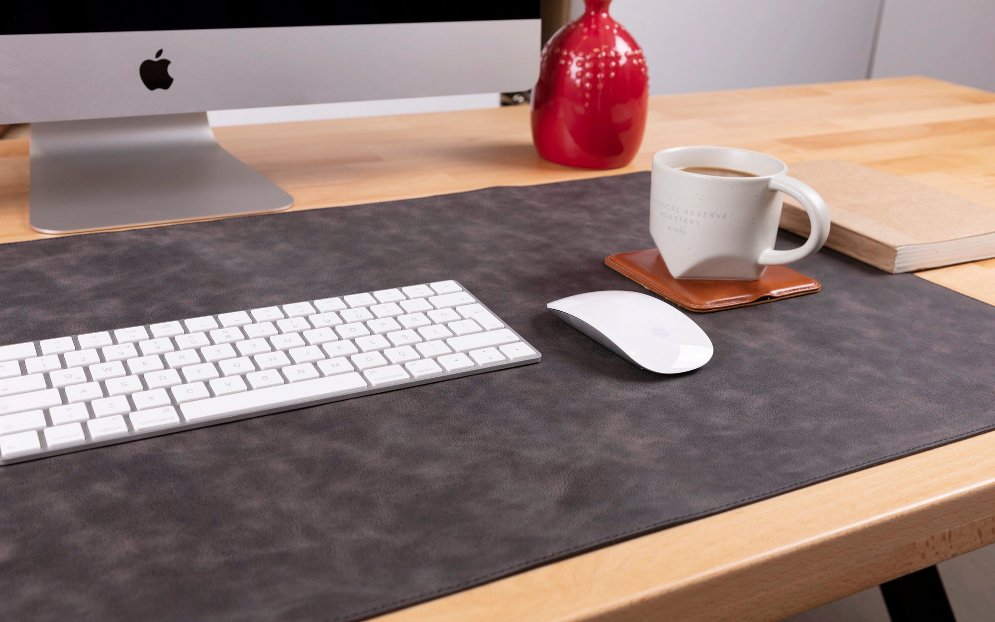 iMac, klavye, fare, kahve kupası, kırmızı vazo ve defter bulunan bir masa düzeni.
