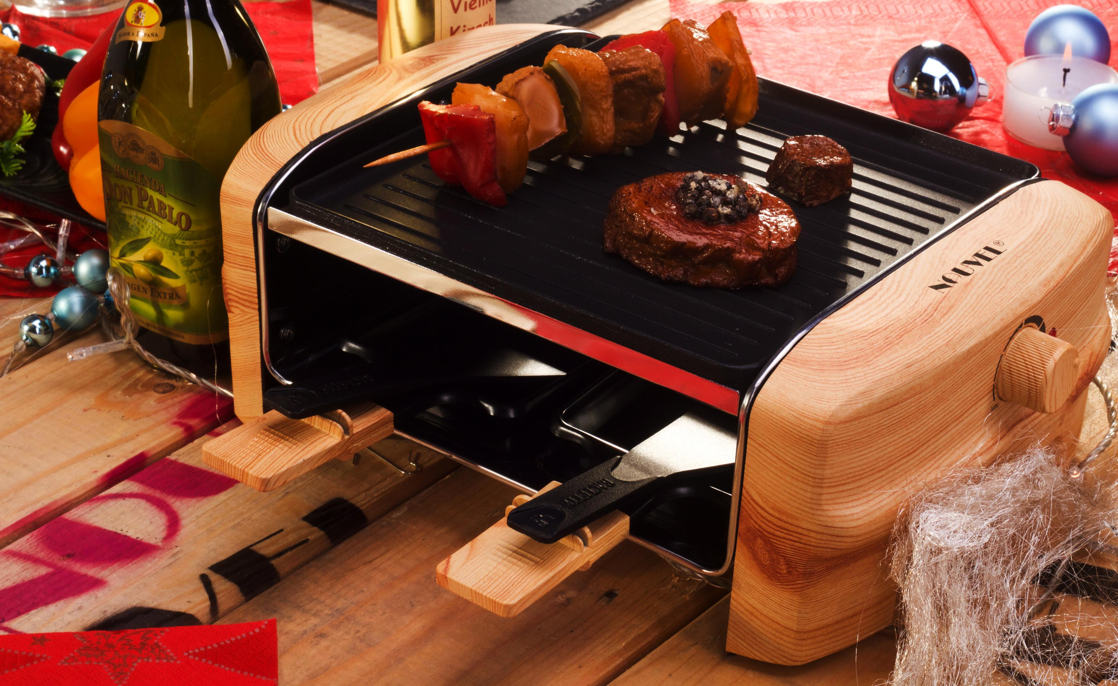 Grill per raclette con carne e spiedini, accanto a una bottiglia d'olio d'oliva e decorazioni.