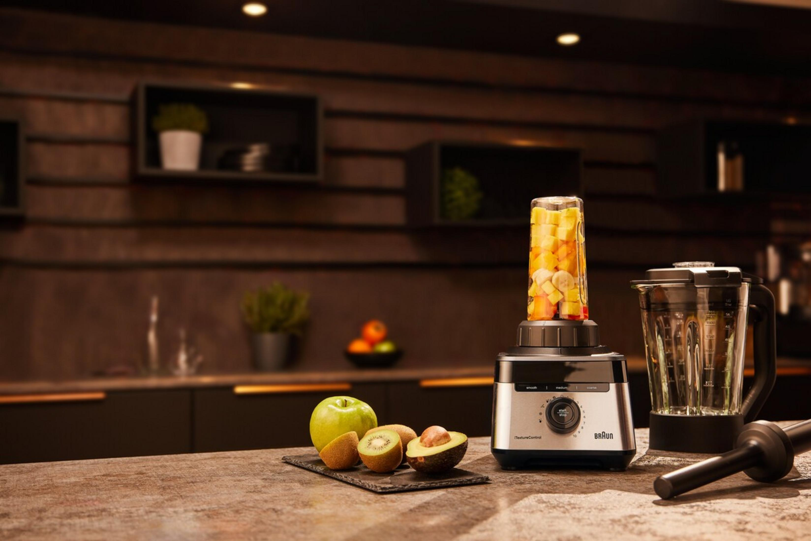 Ein Mixer mit Obst gefüllt, daneben ein Apfel, Kiwi und Avocado auf einem Brett.