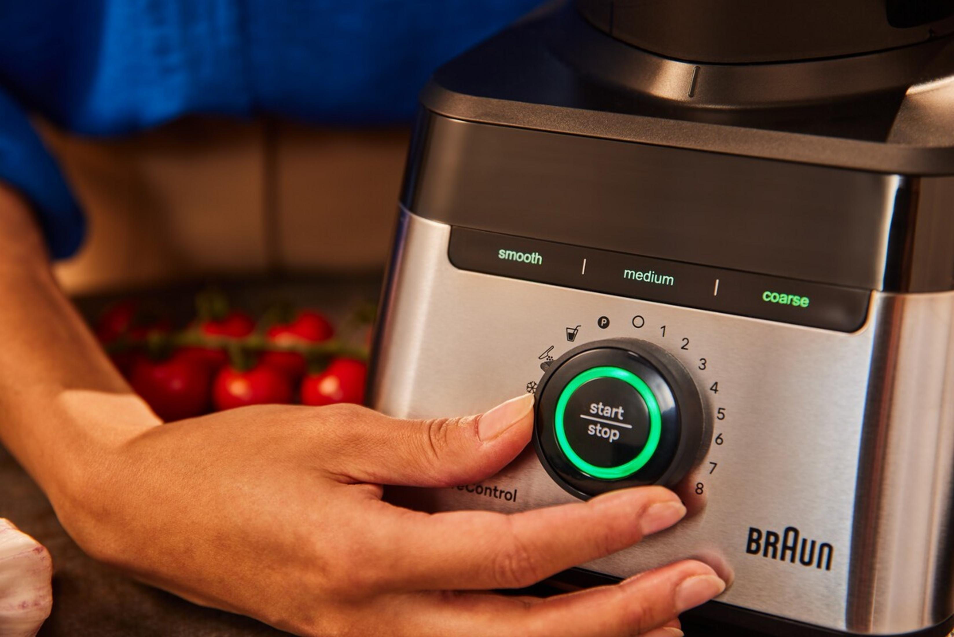 Eine Hand drückt den beleuchteten Start/Stopp-Knopf eines Braun-Mixers mit Tomaten im Hintergrund.