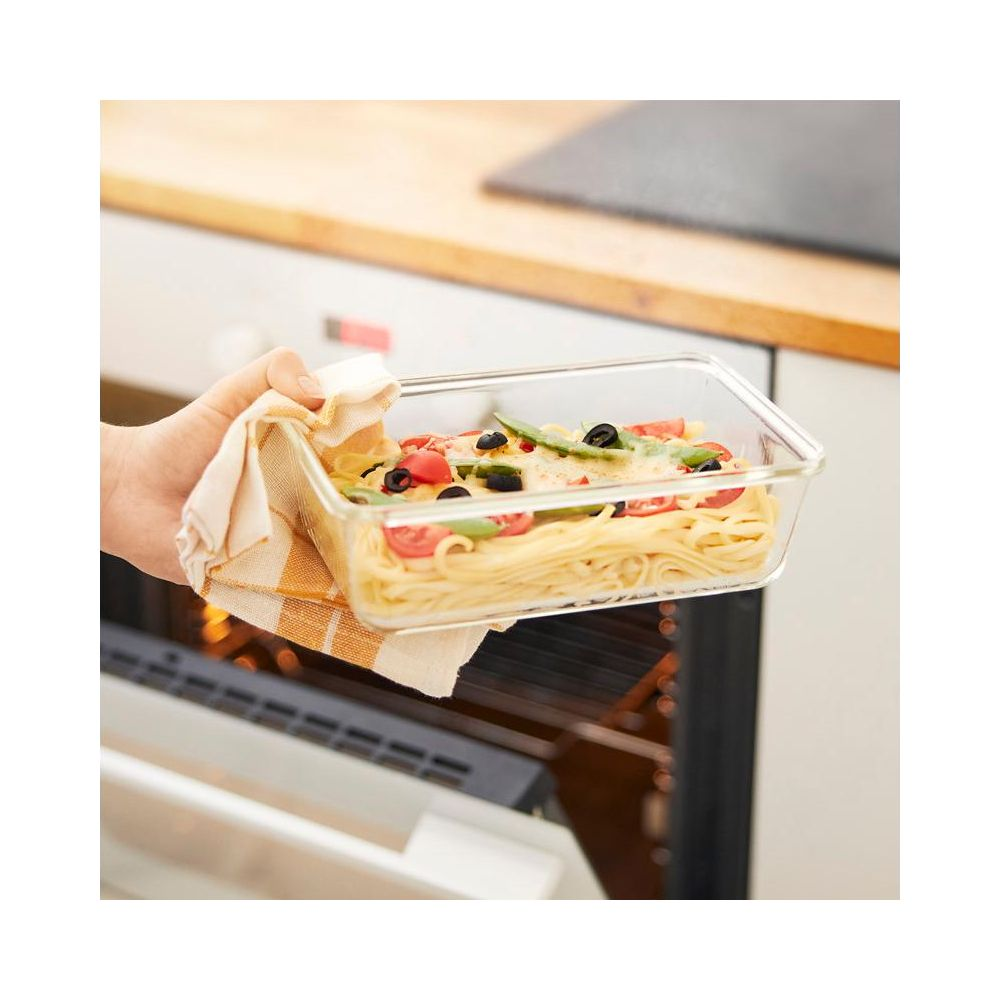 Una mano tira fuori una teglia di vetro con pasta, pomodori e olive da un forno.