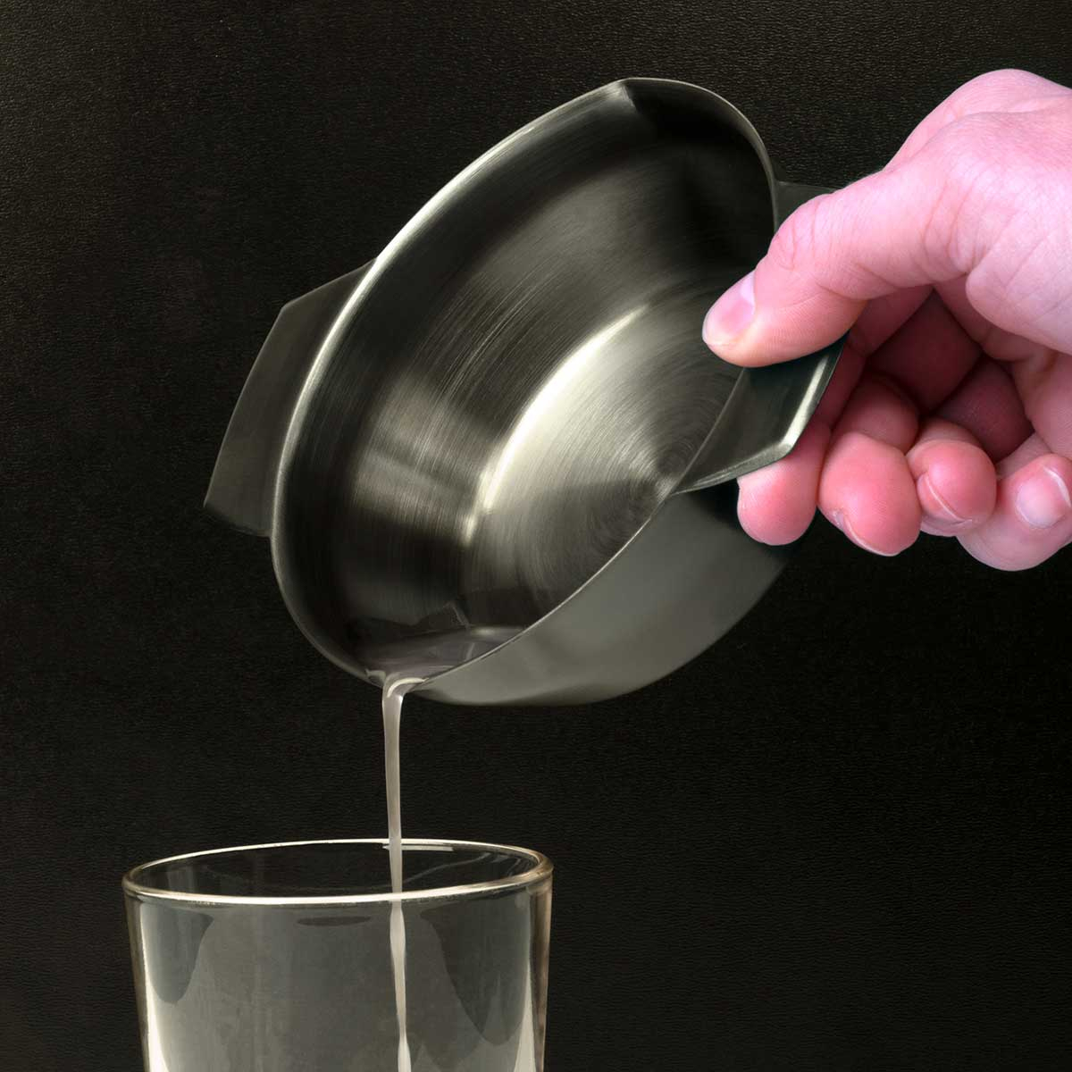Una mano versa un liquido chiaro da una brocca metallica in un bicchiere.
