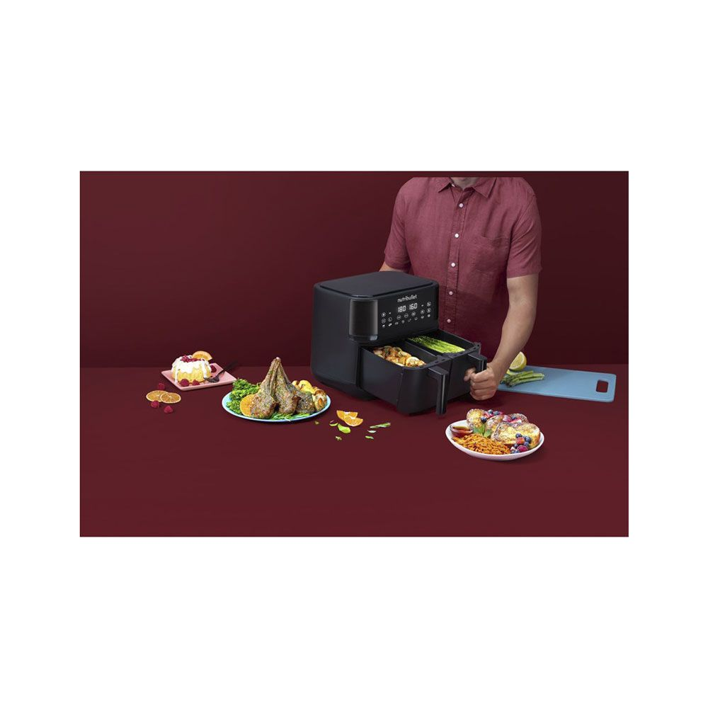 Une friteuse à air noire à double tiroir sur une table avec de la nourriture comme des côtelettes d'agneau et du gâteau.