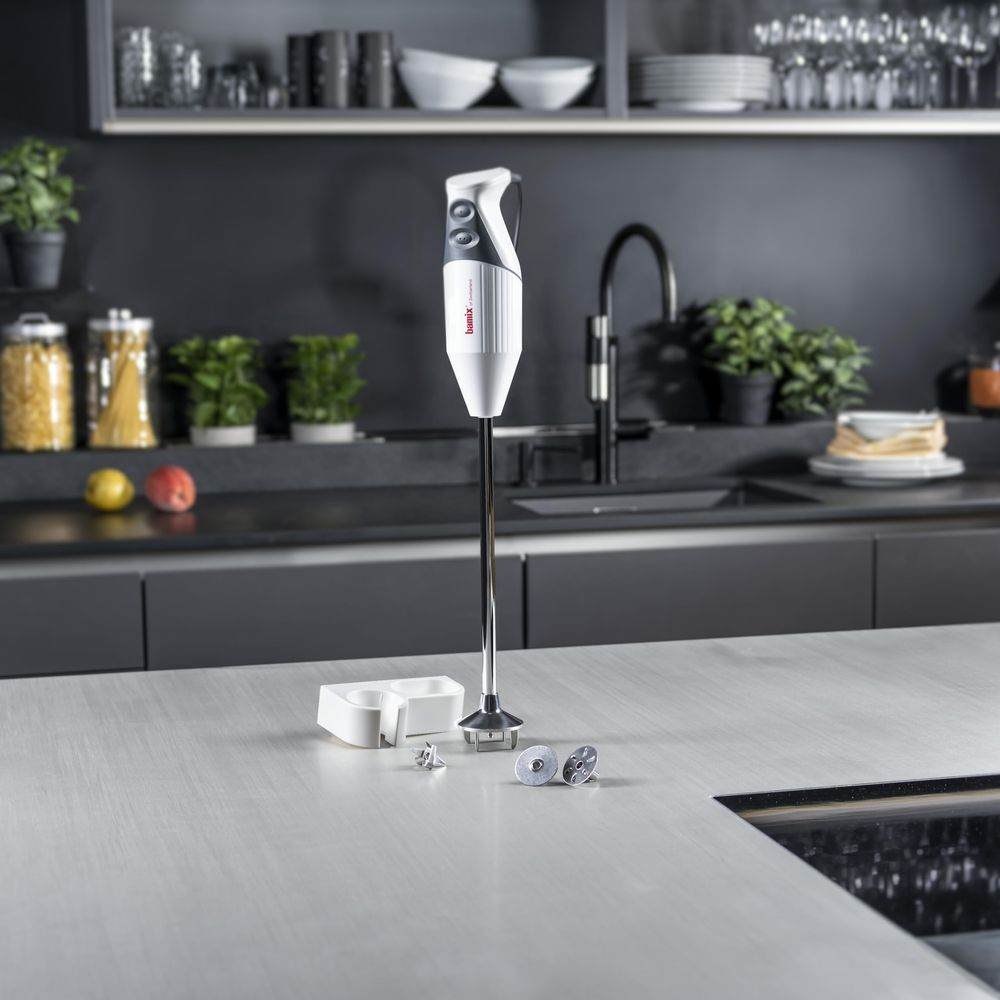 Un mixeur plongeant blanc sur un plan de travail, près d'accessoires et de fruits.