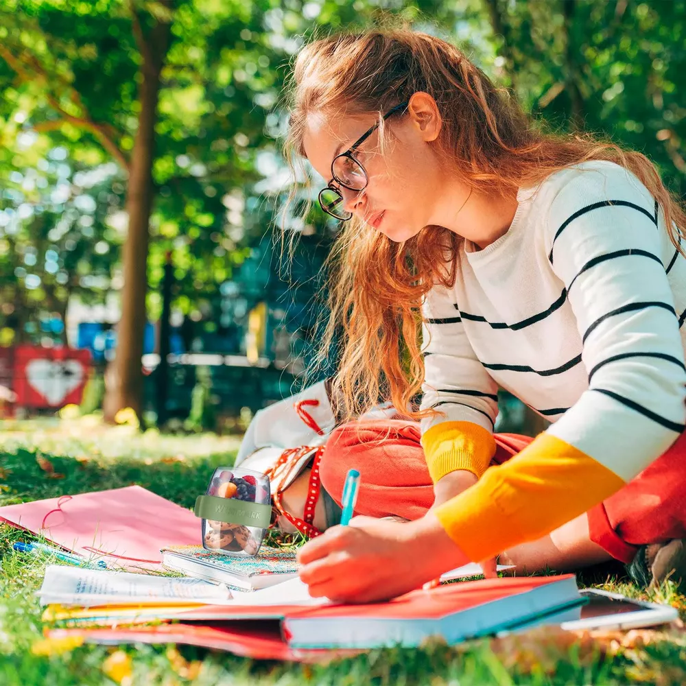 Ein Mädchen mit Brille lernt im Park auf dem Gras mit Büchern und einem Snack.
