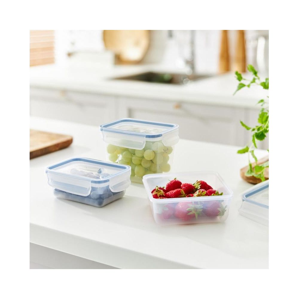 Tre contenitori trasparenti per alimenti con coperchi blu su un piano di lavoro bianco. Uno contiene mirtilli, uno uva, uno fragole.