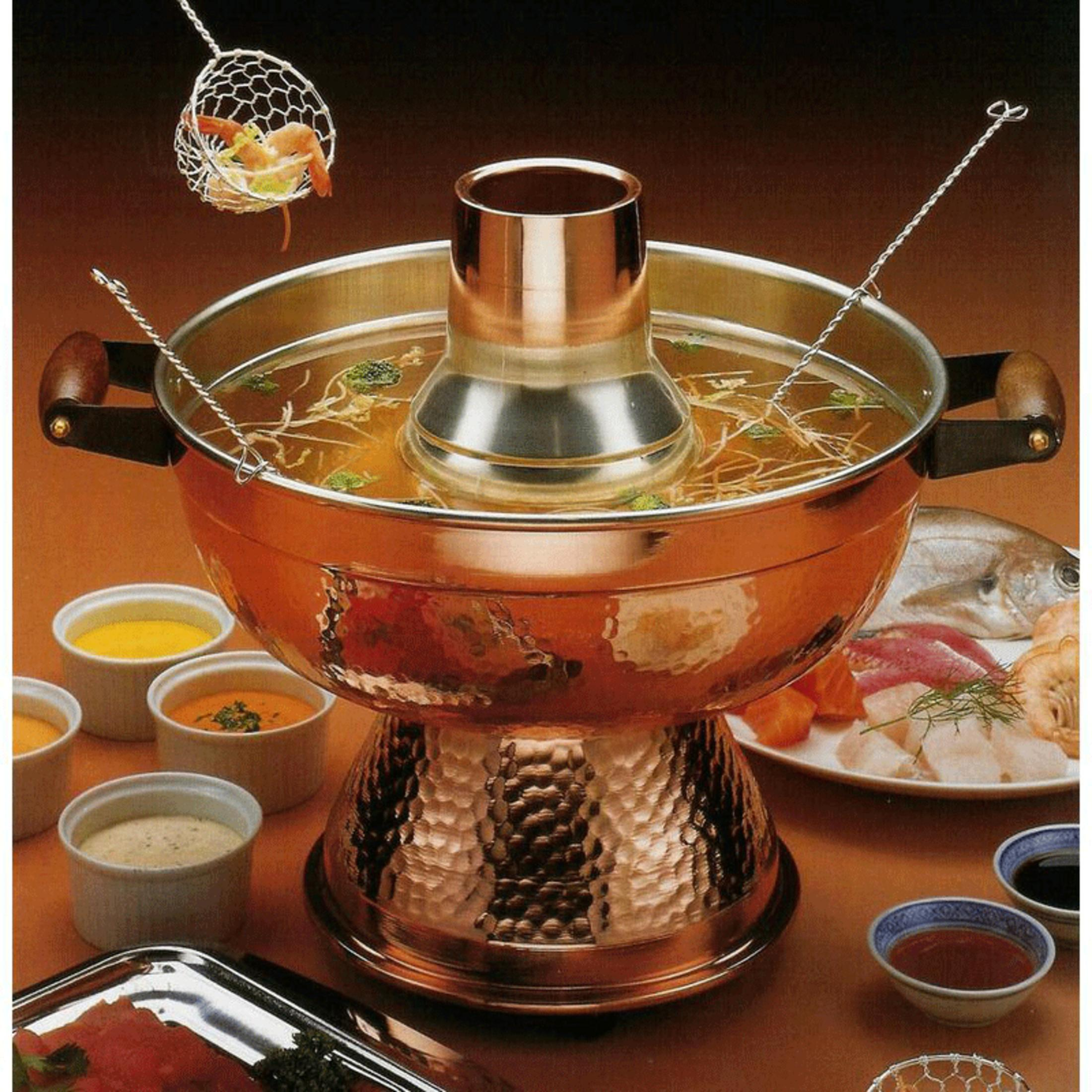 Ein Kupfer-Fondue-Topf mit Brühe, Garnelen, Fisch und Beilagen auf einem Tisch.
