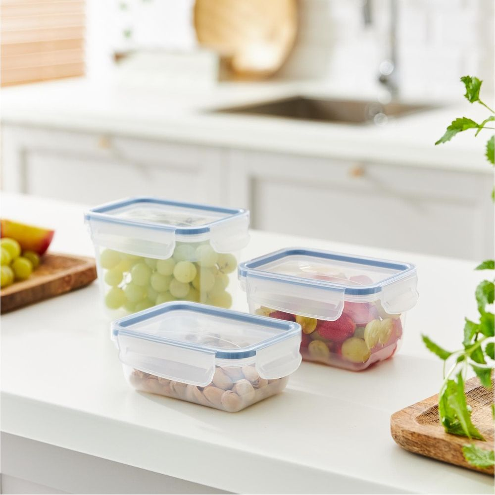 Tre contenitori alimentari in plastica trasparente con coperchi blu su un piano di lavoro bianco, riempiti con uva, noci e bacche.