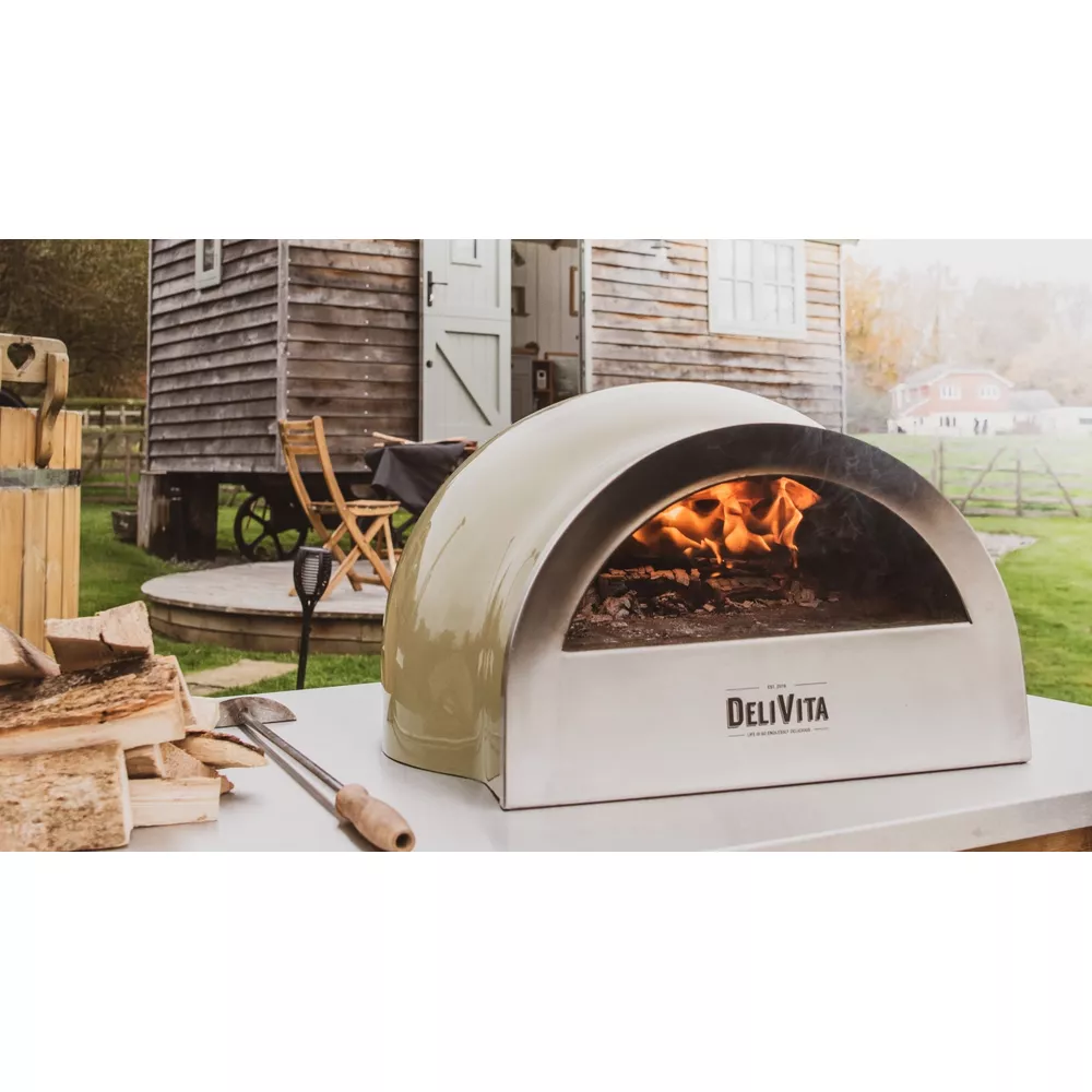 Un four à pizza vert avec un feu allumé est sur une table. Une cabane en bois à l'arrière.