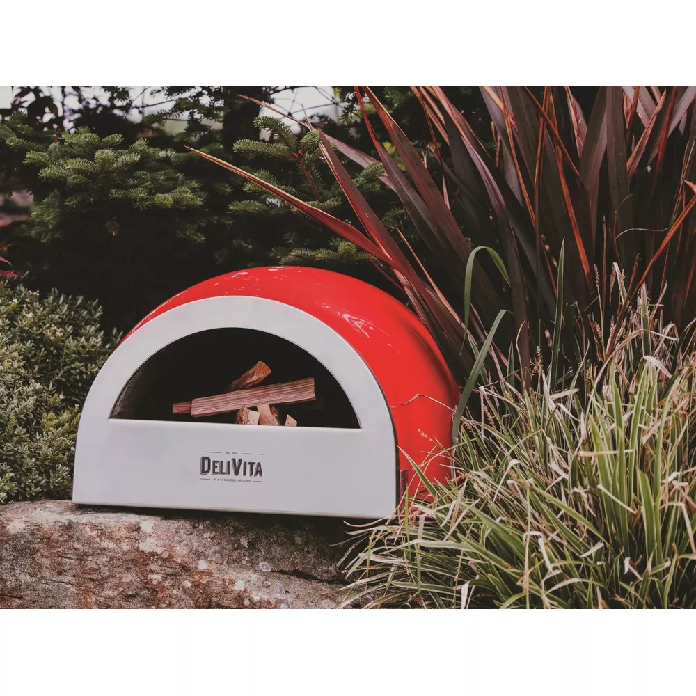 Un four à pizza rouge et blanc avec du bois à l'intérieur, en extérieur parmi la verdure.