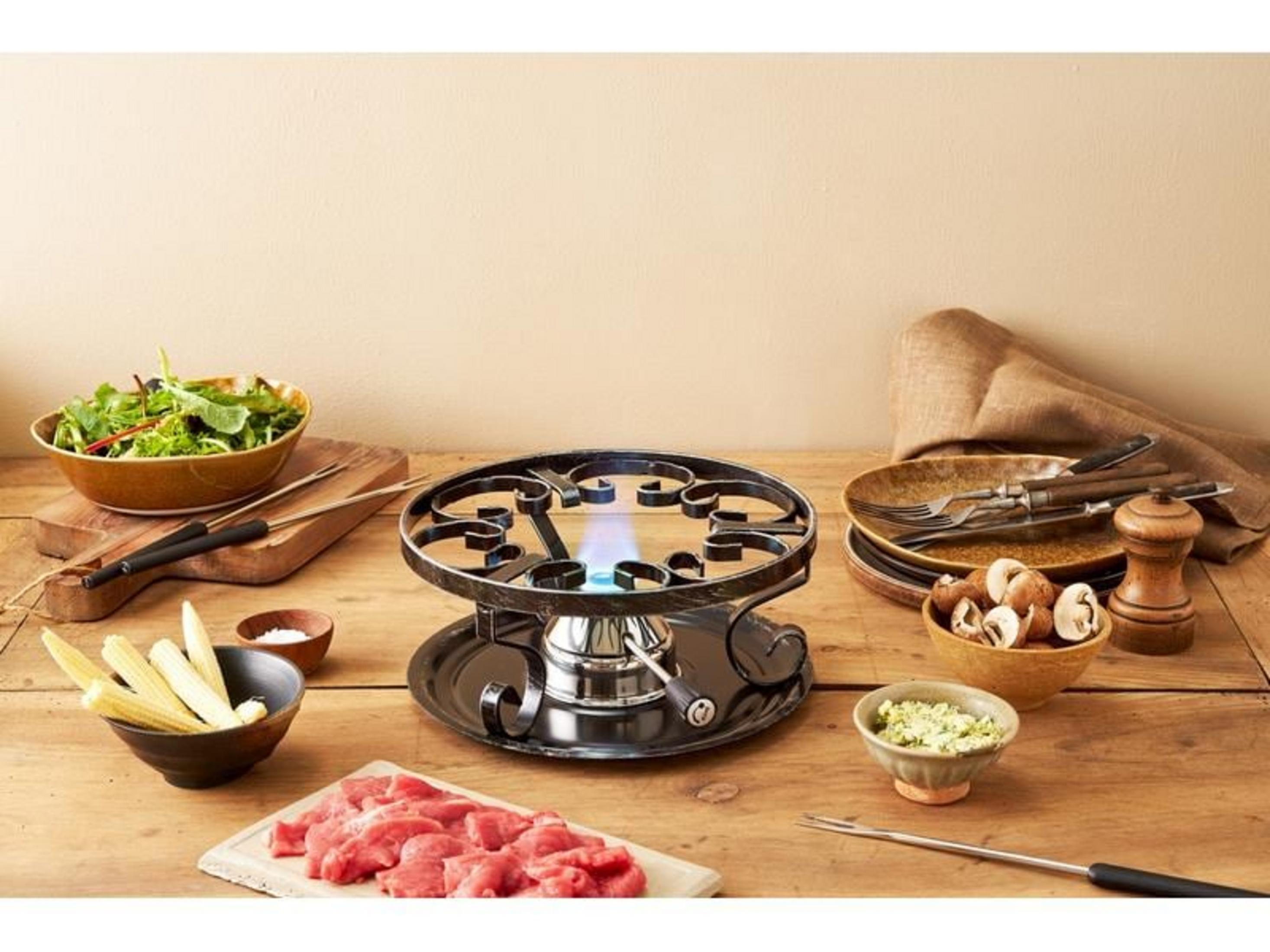 Ein Fondue-Set mit Fleisch, Babykarotten, Salat, Pilzen und Gewürzen auf einem Holztisch.