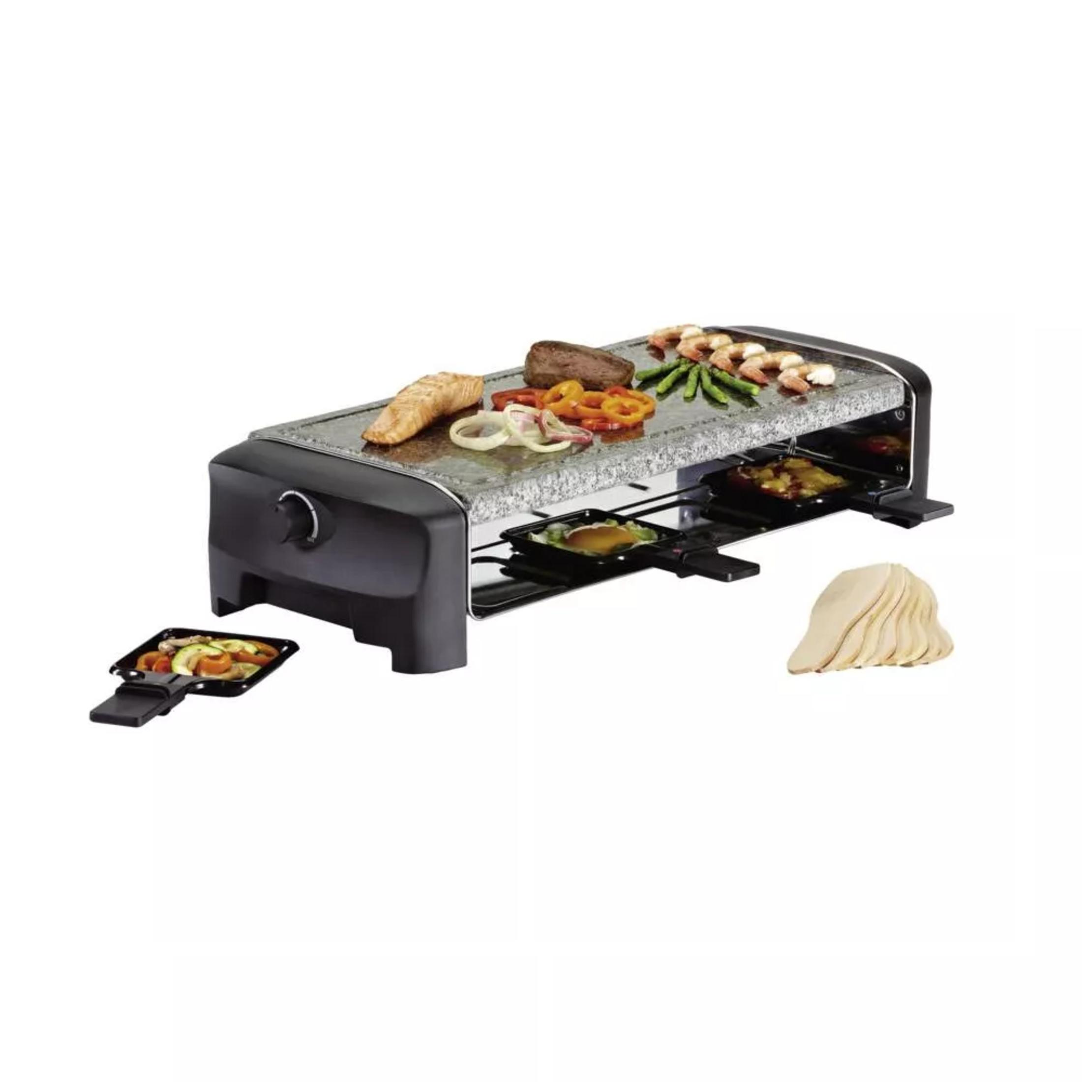 Una griglia raclette nera con piano in pietra è riempita con salmone, bistecca, gamberi, asparagi e verdure, con due padelle raclette sotto e una padella e spatole di legno accanto.