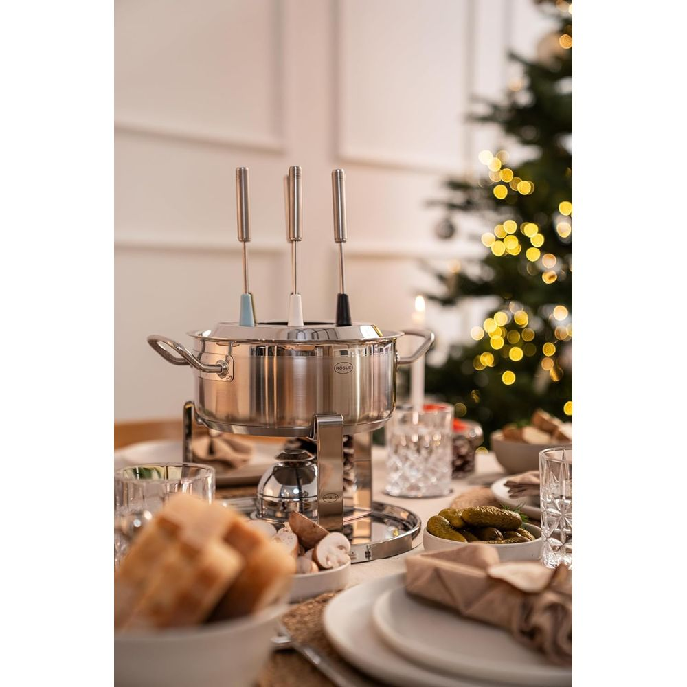 Ein Fondue-Set aus Edelstahl mit drei Gabeln auf einem Esstisch, verschwommene Weihnachtsbaumlichter im Hintergrund.