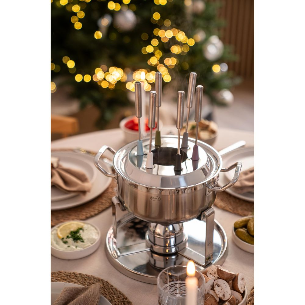 Ein Fondue-Set aus Edelstahl mit bunten Gabeln auf einem Tisch, einer brennenden Kerze und einem verschwommenen Weihnachtsbaum im Hintergrund.
