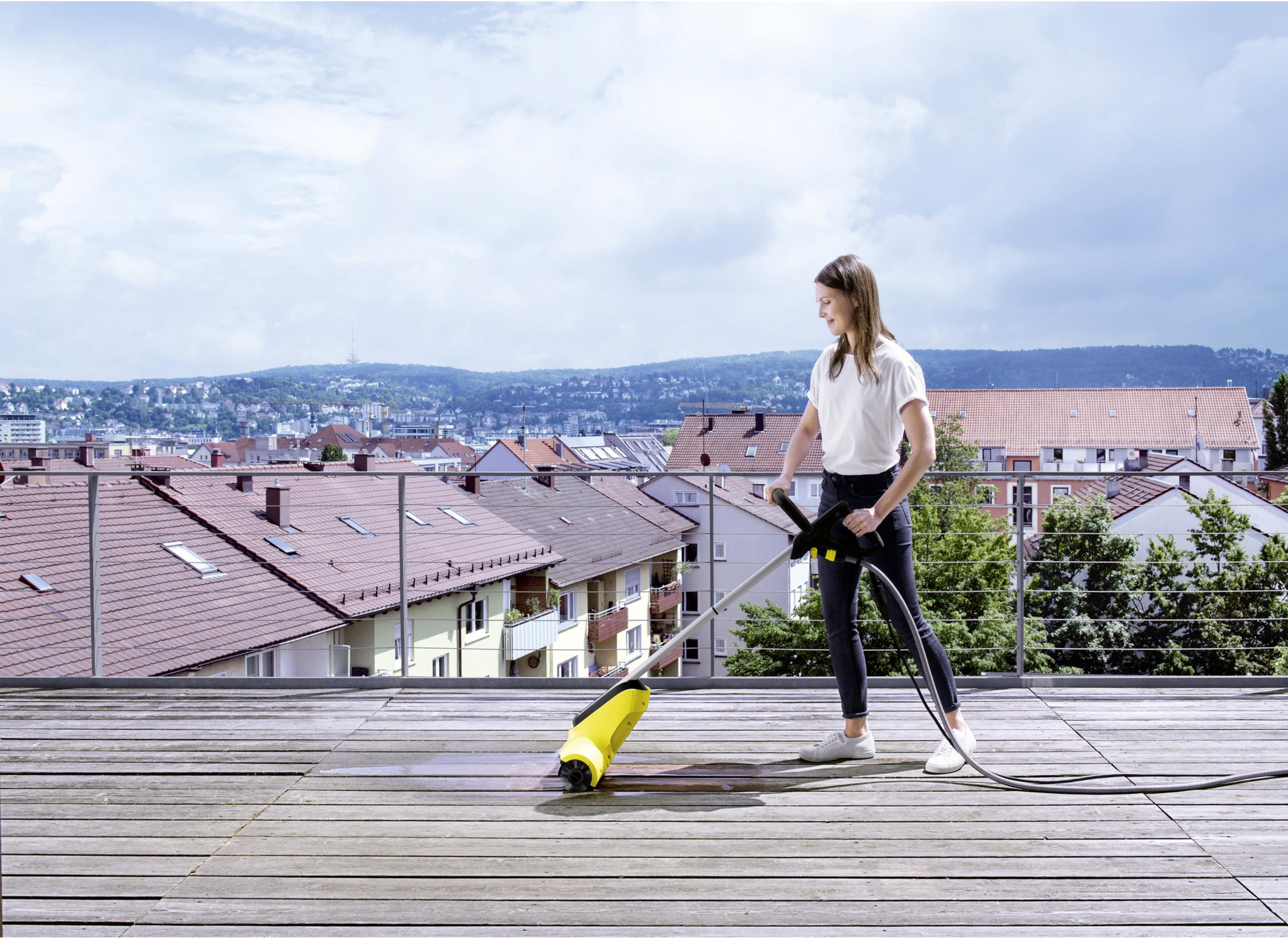 Frau reinigt Holzdeck mit Hochdruckreiniger, Stadtblick im Hintergrund.
