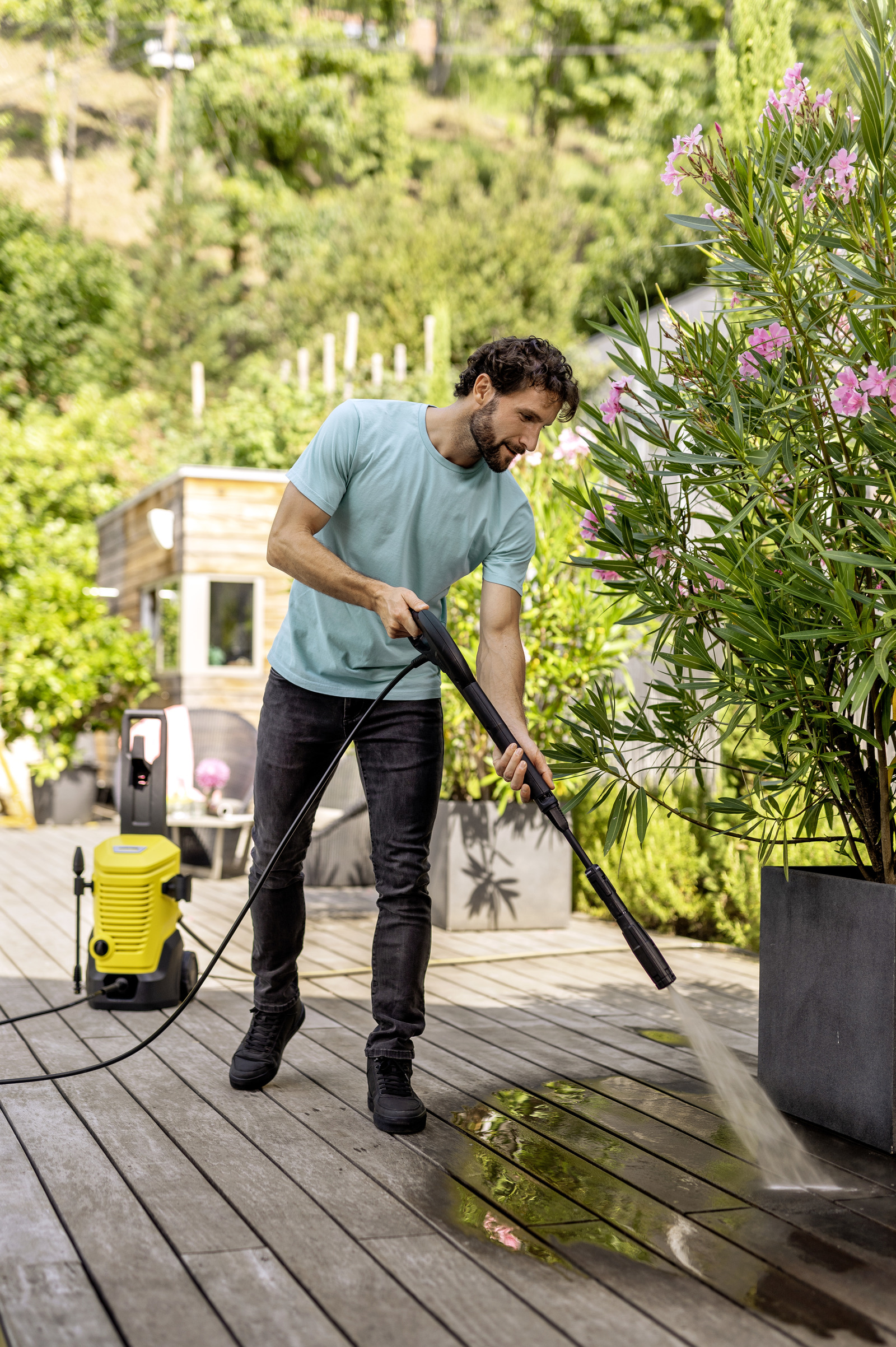 Mężczyzna czyści drewniany taras myjką ciśnieniową obok donicy z różowymi kwiatami.