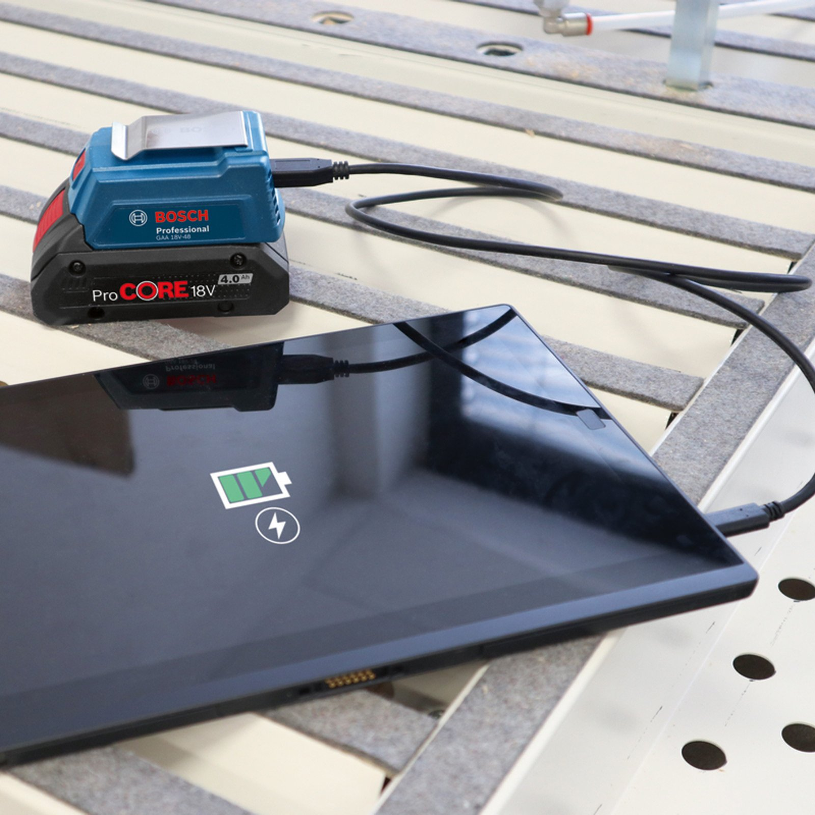Bir Bosch pil, dokulu bir yüzeyde USB kablosuyla bir tableti şarj ediyor.