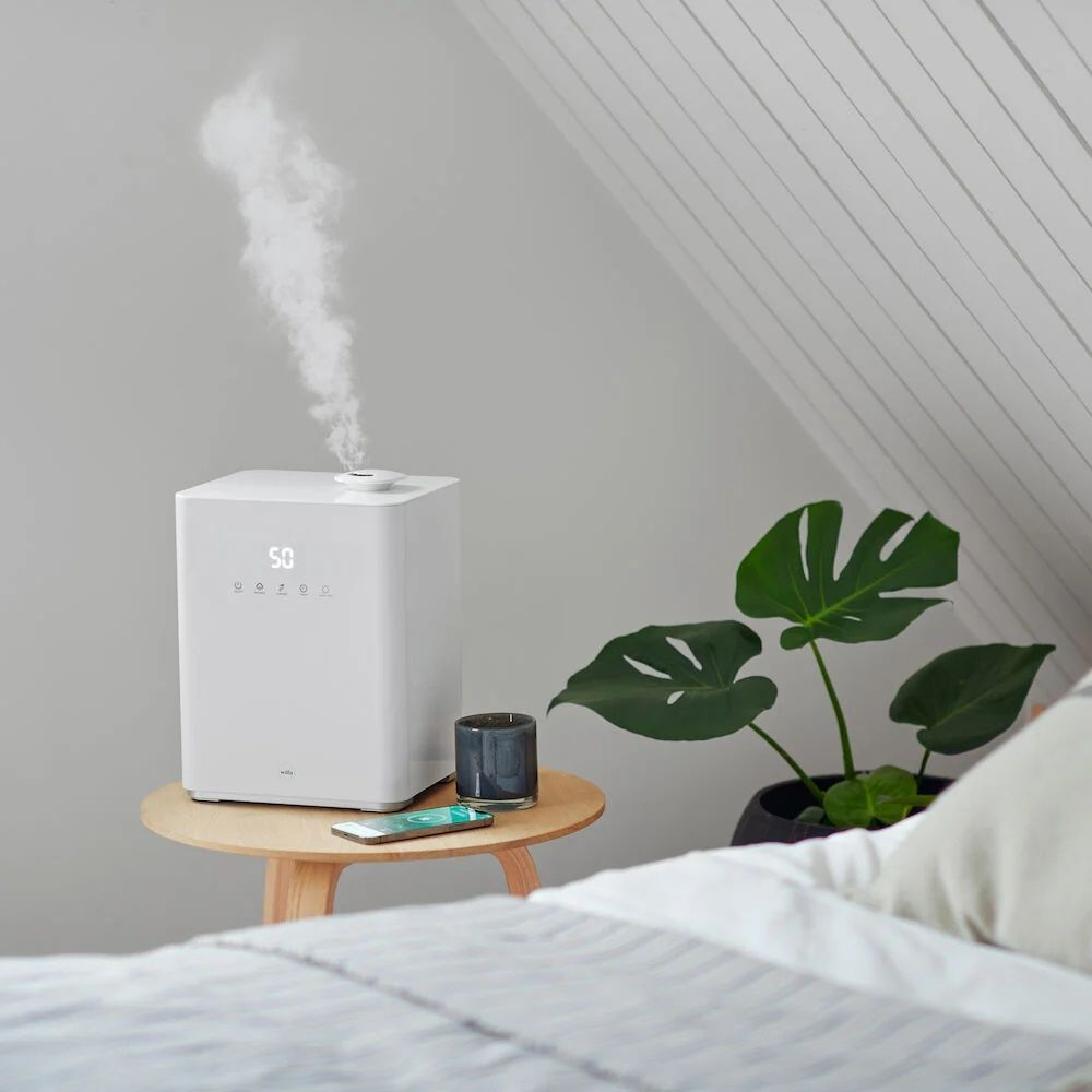 Un umidificatore bianco emette vapore, accanto a una pianta e un telefono.