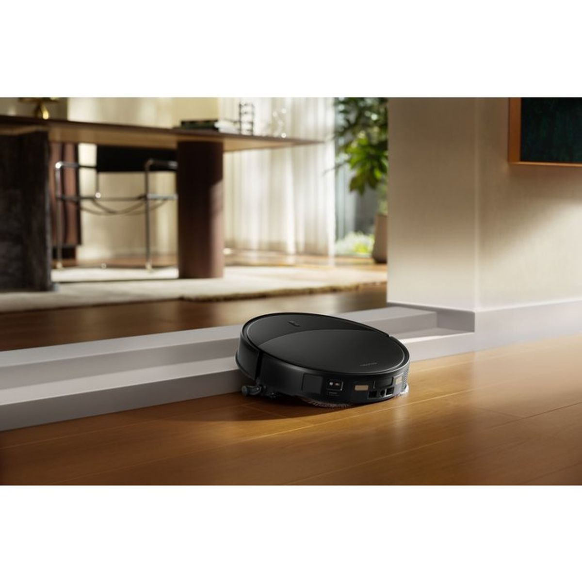 Un robot aspirador negro limpia un suelo de madera en un salón moderno.