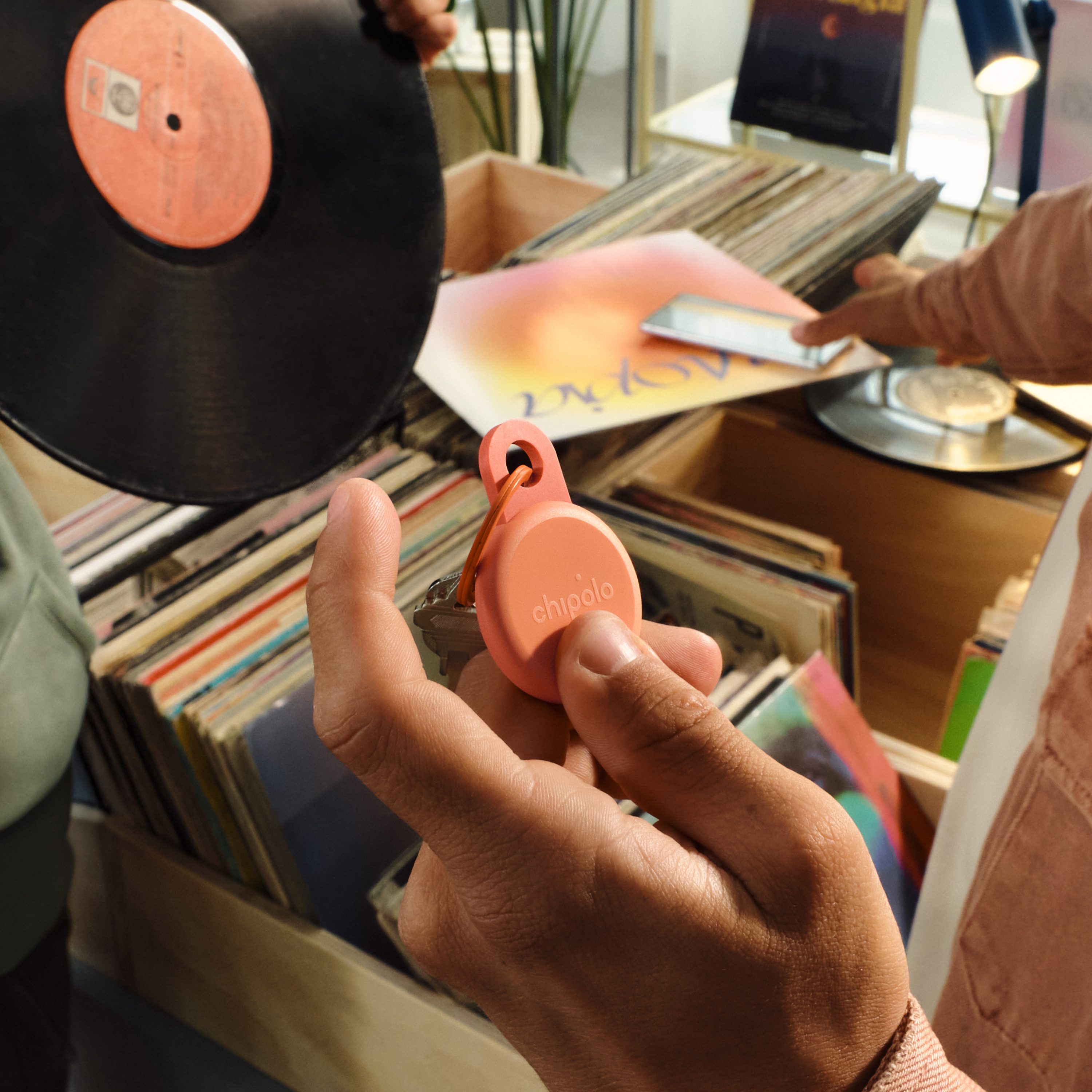 A hand holds an orange Chipolo keychain in front of a collection of vinyl records.