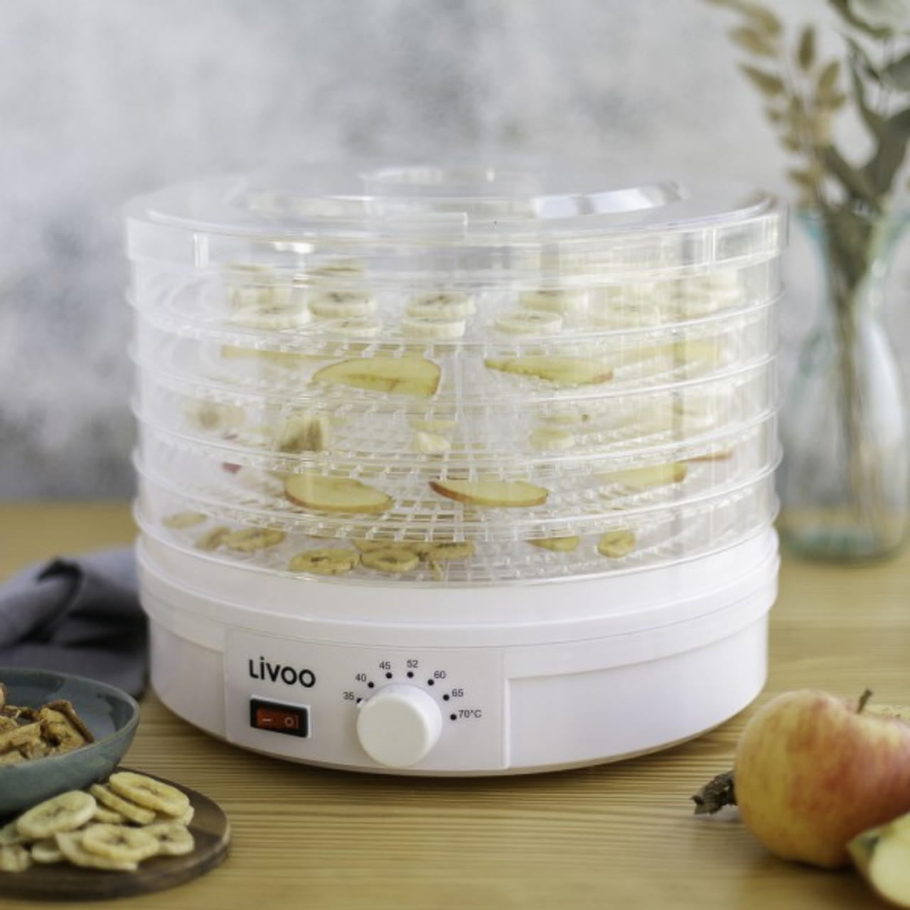 Ein weißer Dörrautomat mit Obst auf mehreren Ebenen, daneben frische Äpfel.